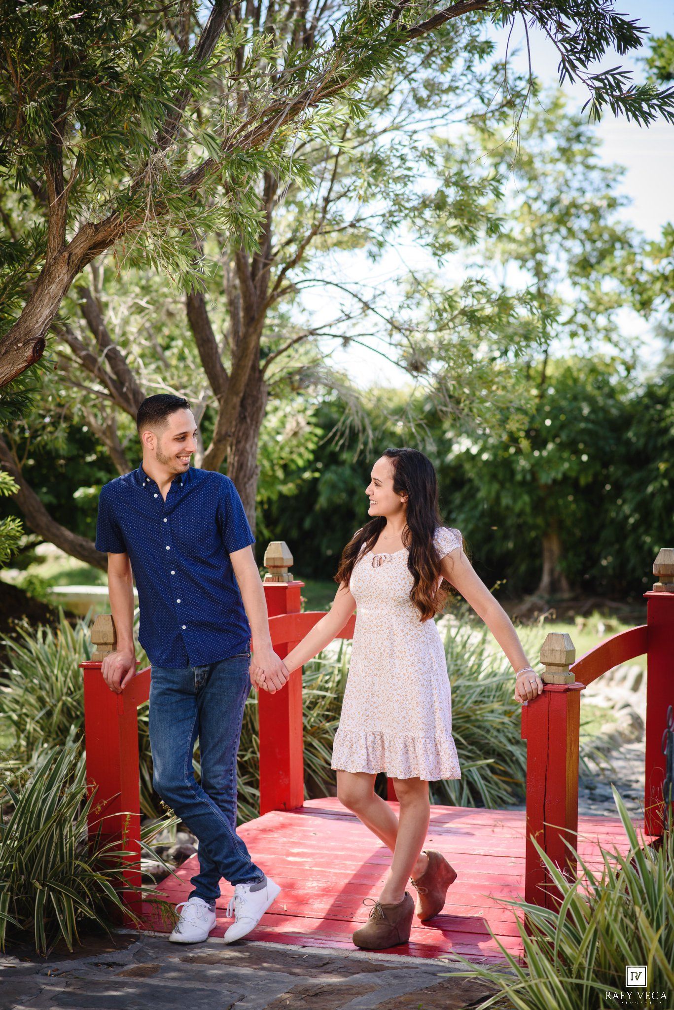 Couple holding hands on a red bridge in a lush garden, man in blue shirt and jeans, woman in white dress.