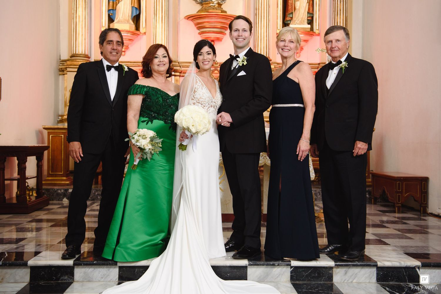 Wedding party posing in church, bride in white gown, groom in tuxedo, flanked by family in green and navy formal wear.