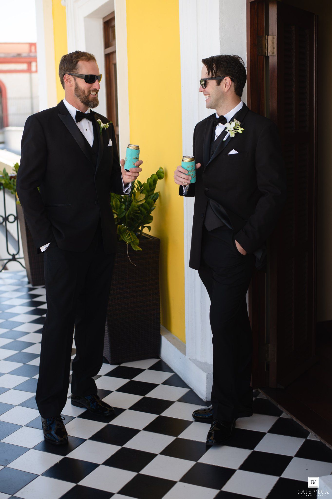 Two groomsmen in black tuxedos with boutonnieres holding drinks on a checkered floor by a yellow wall.