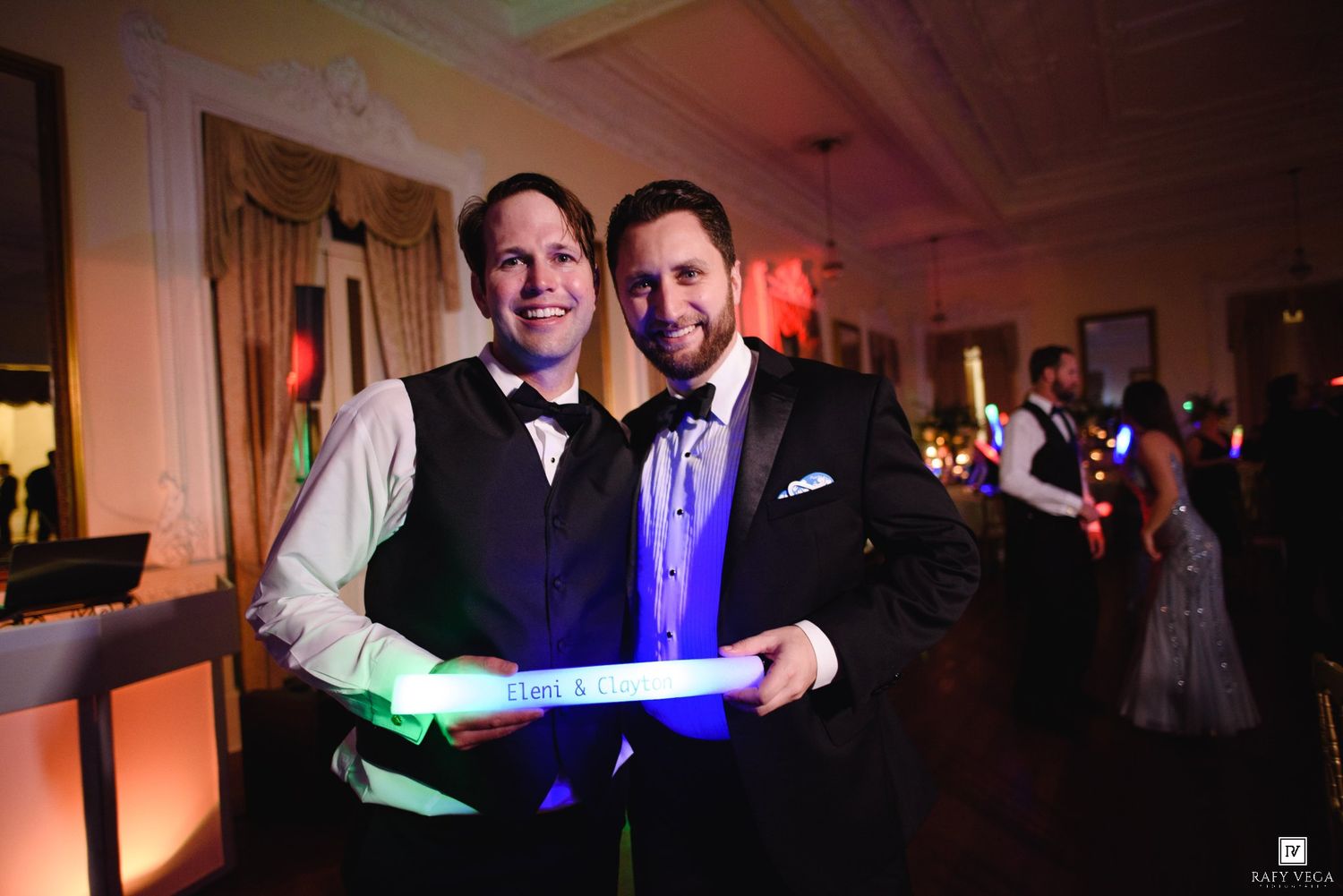 Two men in formal attire smile holding glow sticks at an elegant wedding reception.