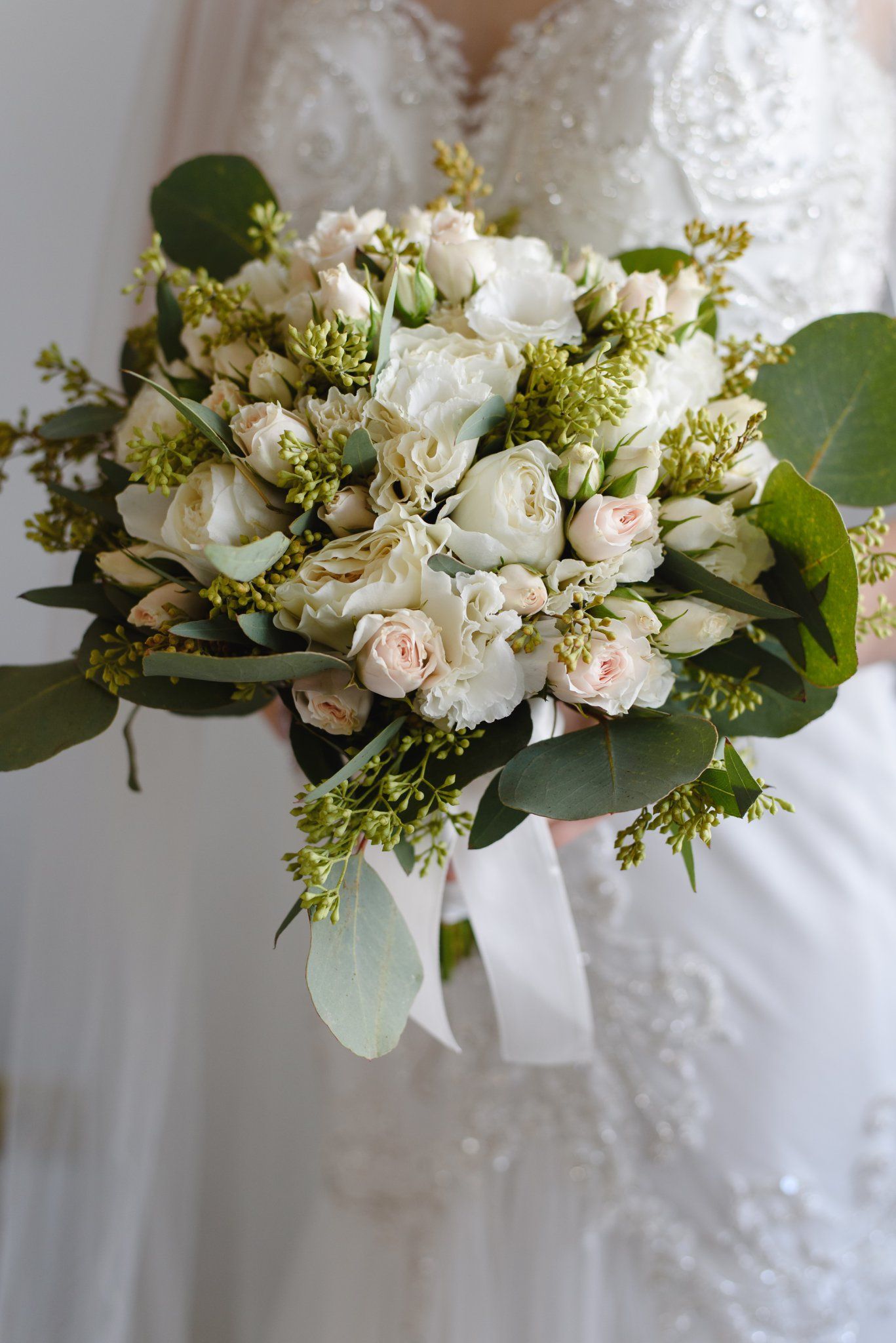 Close-up of elegant bridal bouquet with white roses, blush blooms, and green eucalyptus leaves tied with white ribbon.