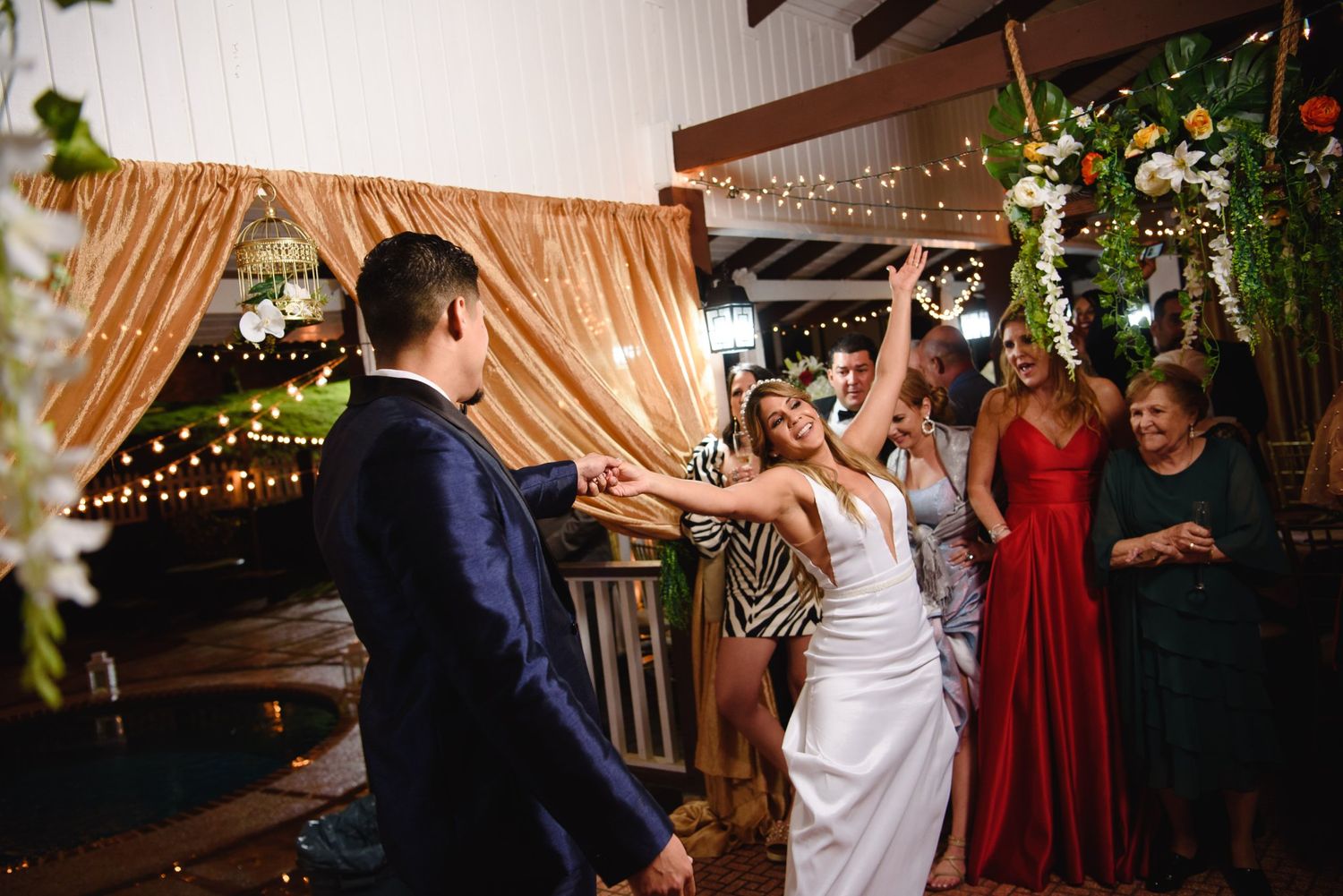 Bride and groom dancing joyfully with guests at elegant wedding reception with string lights and gold drape backdrop.