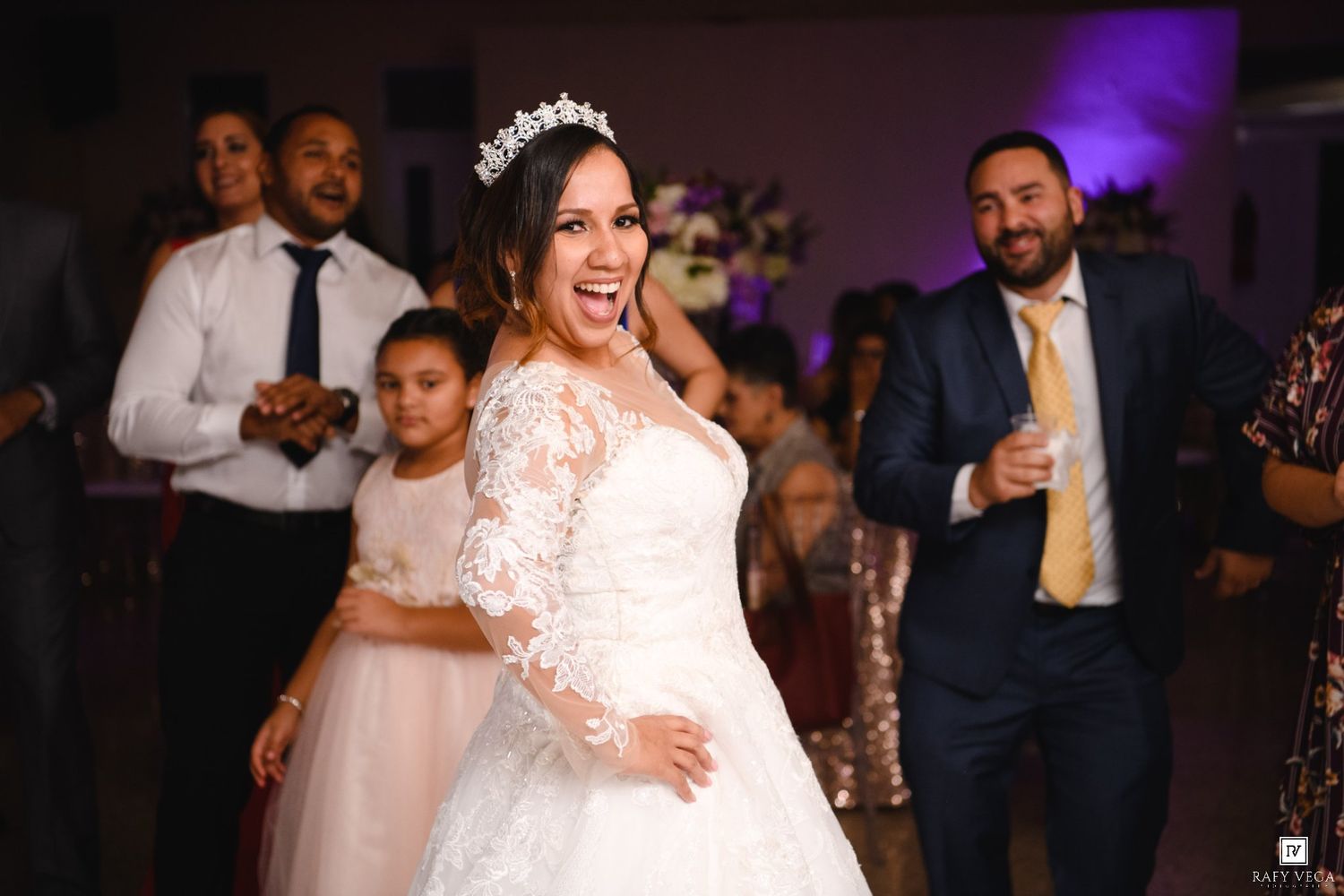 Bride in lace wedding gown and tiara laughing joyfully on dance floor surrounded by guests at wedding reception.