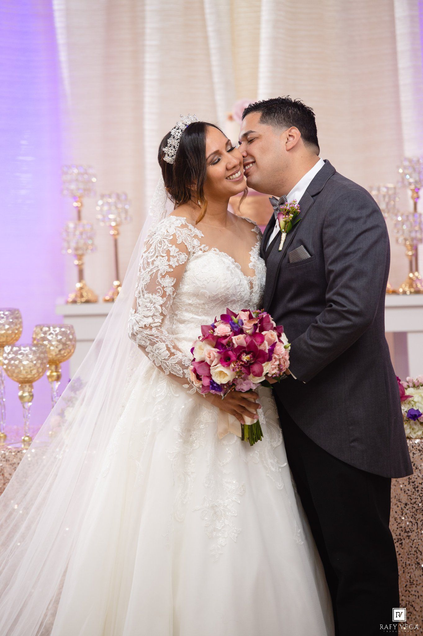 Bride and groom share a romantic kiss at elegant wedding reception with floral bouquet and gold candelabras.