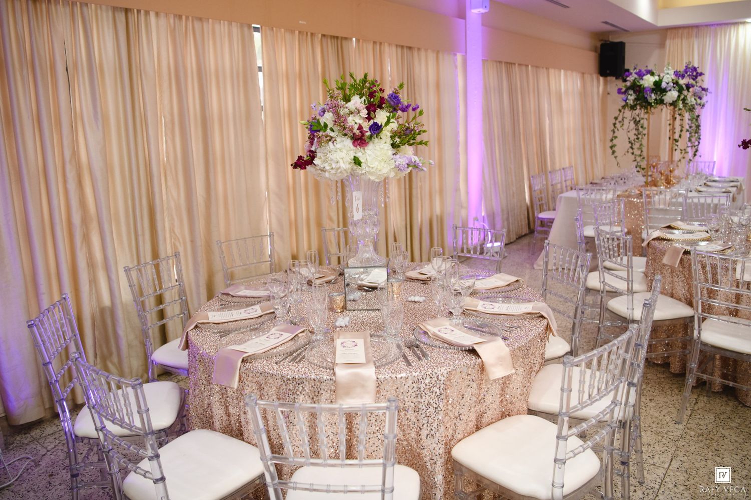 Elegant wedding reception venue with round tables, gold lace linens, chiavari chairs, and tall floral centerpieces under purple uplighting.