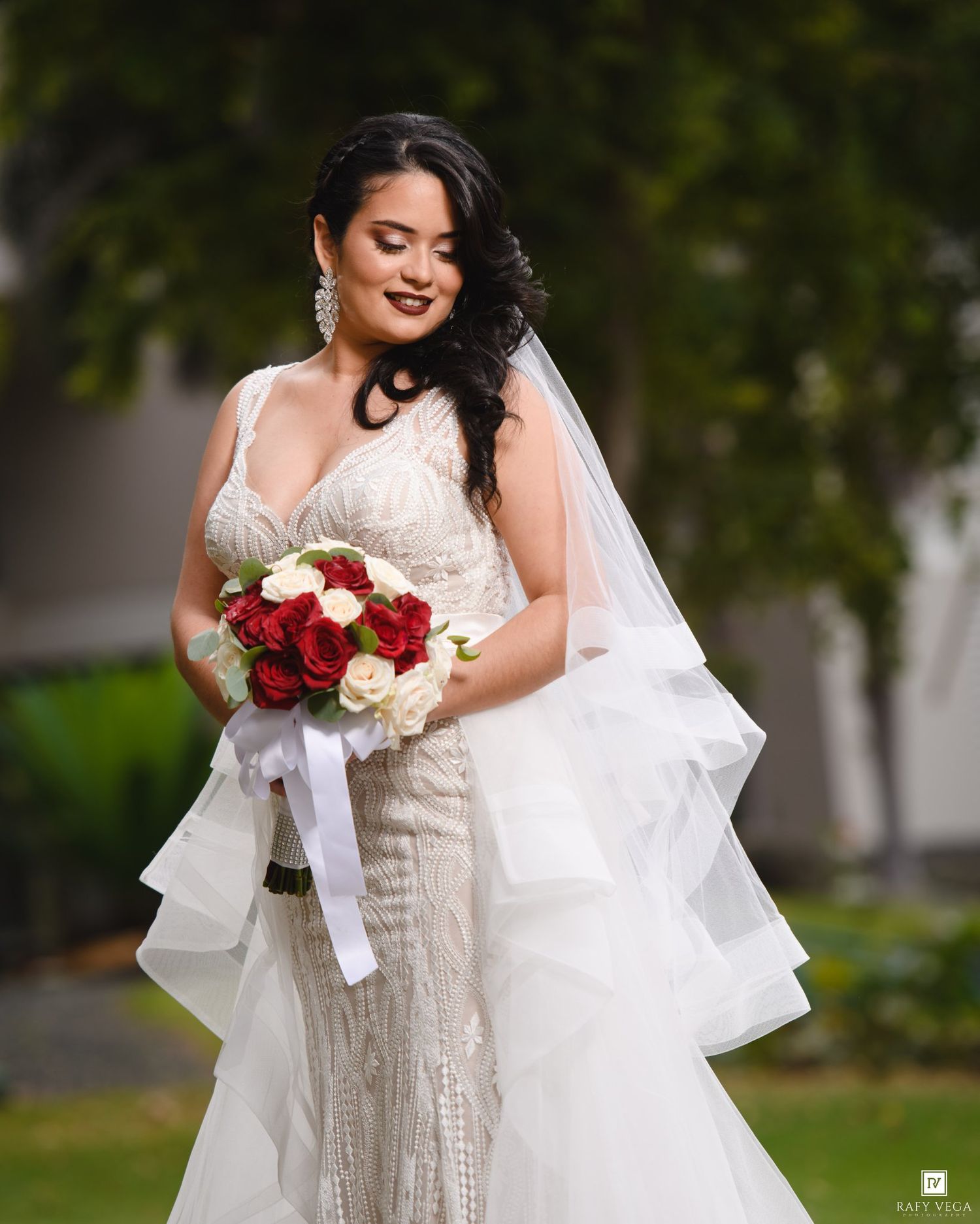 Bride in beaded mermaid gown holding red and white rose bouquet with layered veil outdoors.
