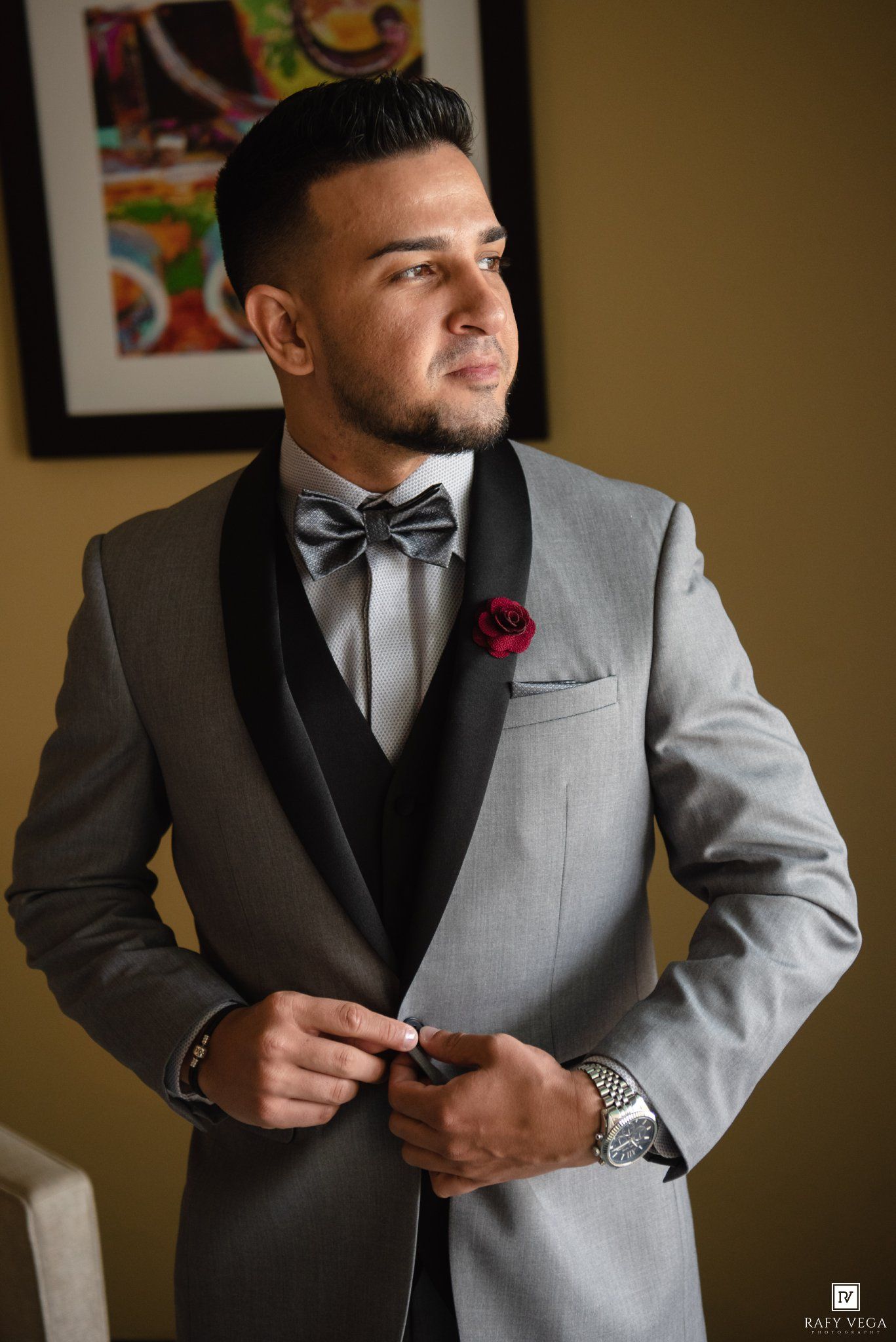 Groom in gray tuxedo with black lapels, silver bow tie, and red rose boutonniere buttoning his jacket.