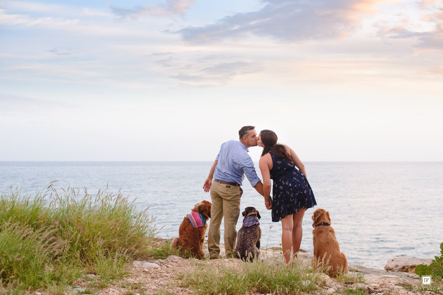 Couple kissing with three dogs by ocean shore at sunset on rocky coastline.