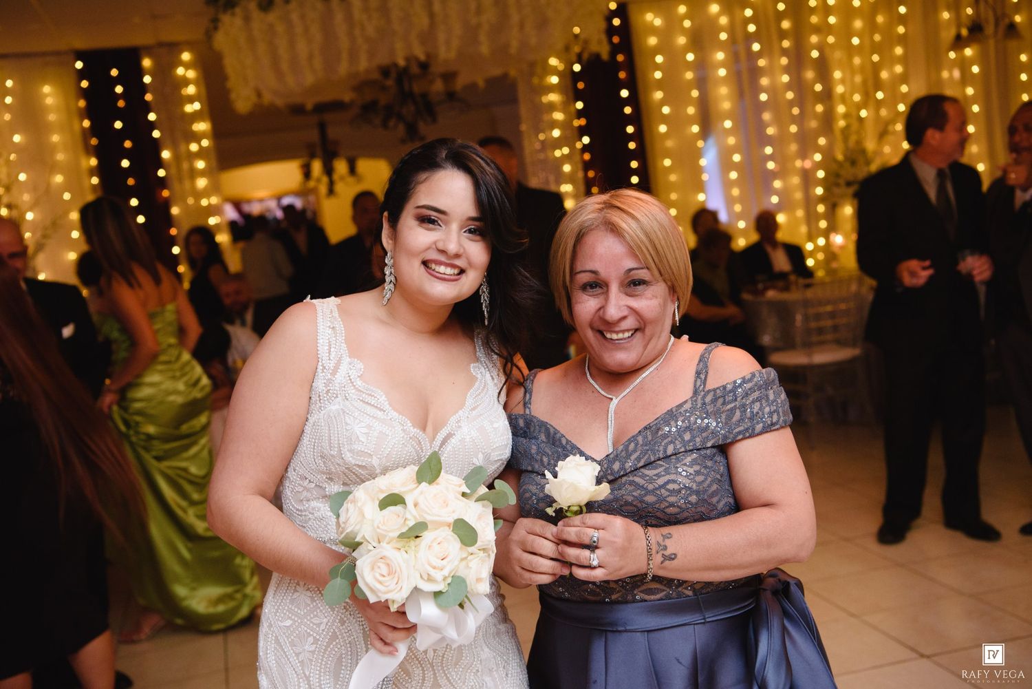 Bride in lace gown holding white bouquet poses with older woman in blue dress at elegant wedding reception.