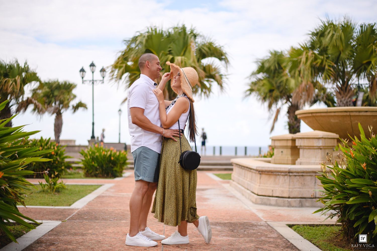 Couple embracing and kissing near a fountain with palm trees in a sunny tropical plaza.