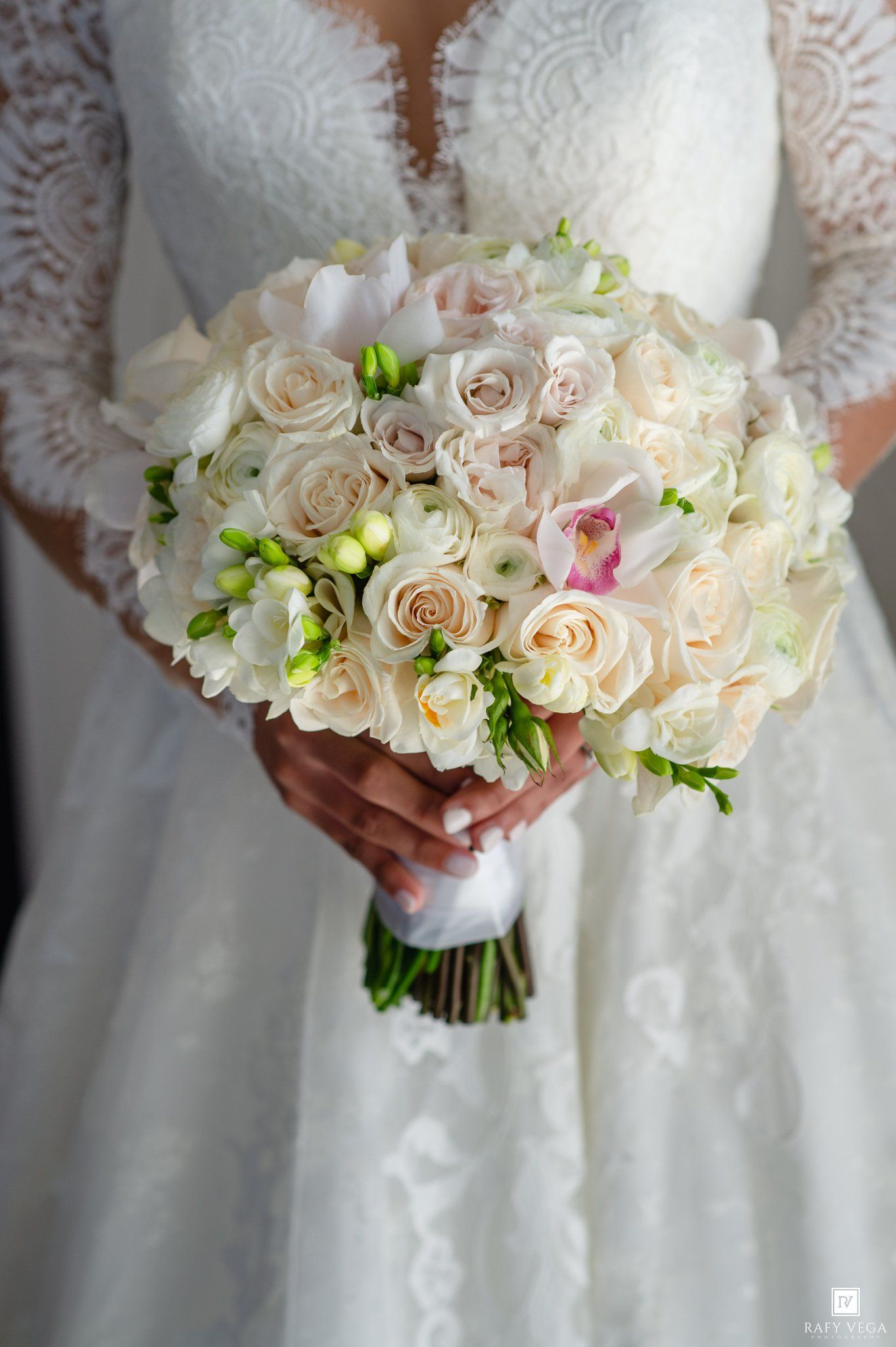 Bride holding elegant white and blush bridal bouquet with roses, orchids, and freesias against lace wedding gown.