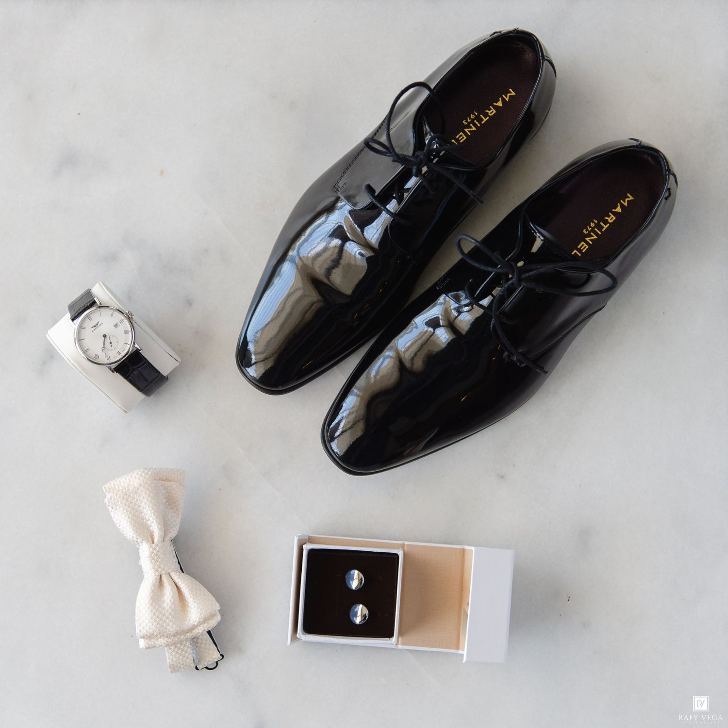 Black patent leather dress shoes with cufflinks, bow tie, and watch arranged on marble surface for a formal wedding ensemble.