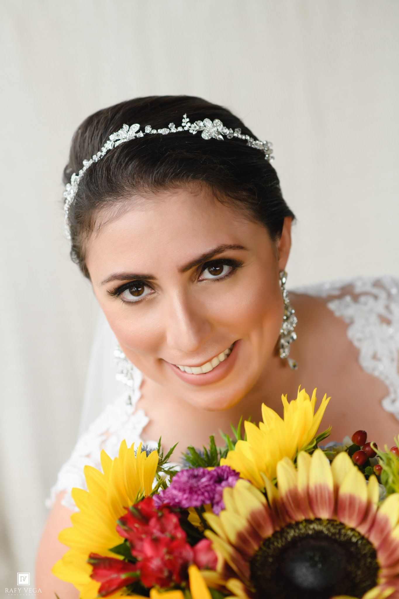 Smiling bride wearing a crystal tiara and lace gown holds a vibrant sunflower bouquet with red and purple blooms.