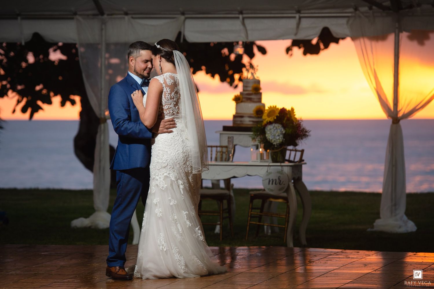 Bride and groom share romantic dance at sunset wedding reception under elegant tent with tiered cake backdrop.