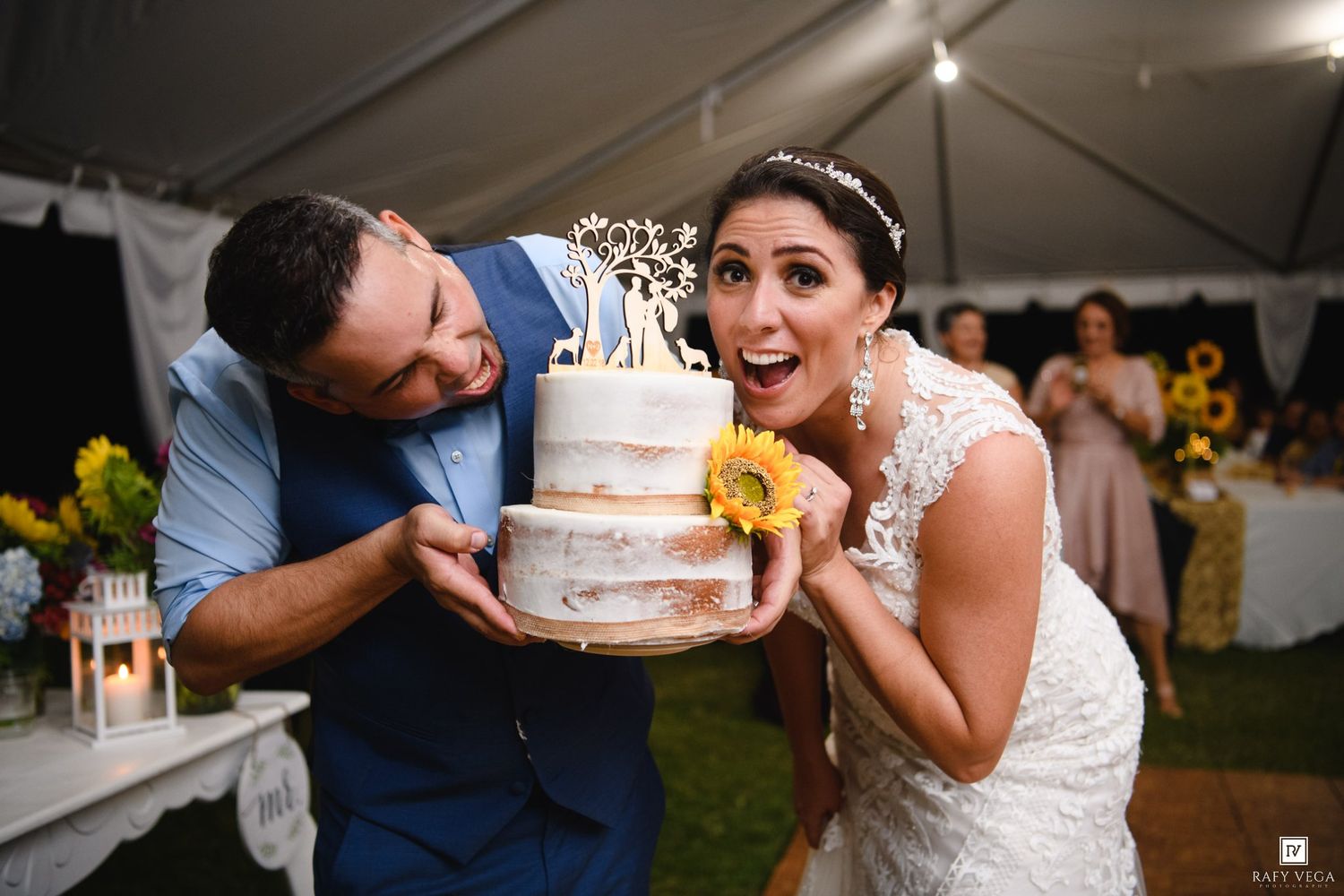 Bride and groom joyfully holding a rustic wedding cake topped with sunflowers and a couple topper at their outdoor reception.