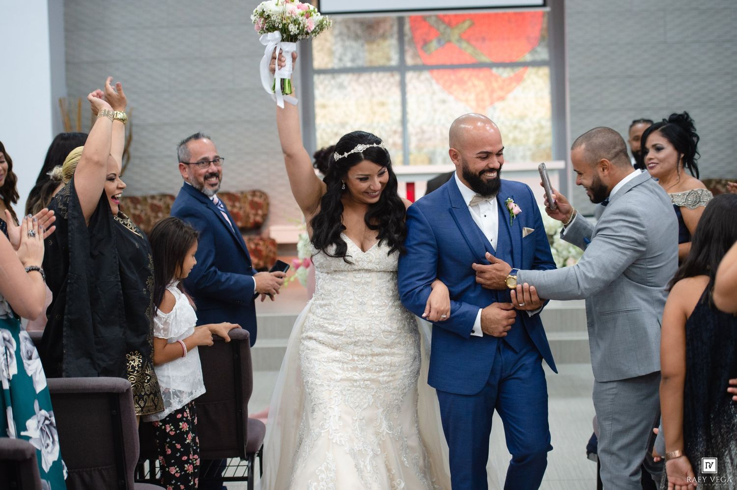 Joyful newlyweds walk down the aisle after wedding ceremony, bride in lace gown holding bouquet aloft.