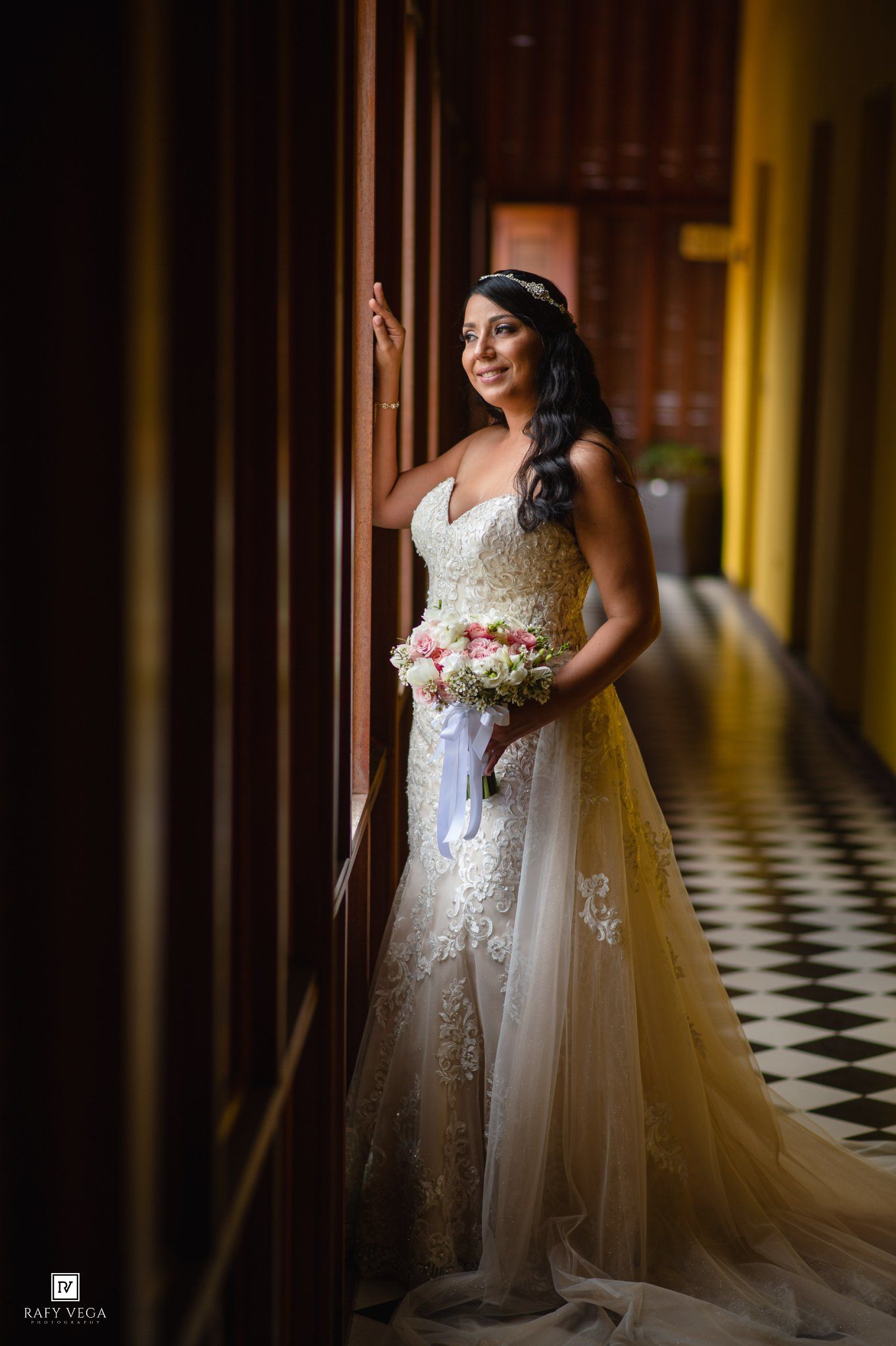 Bride in lace wedding gown holding floral bouquet, standing in elegant hallway with checkered floor.