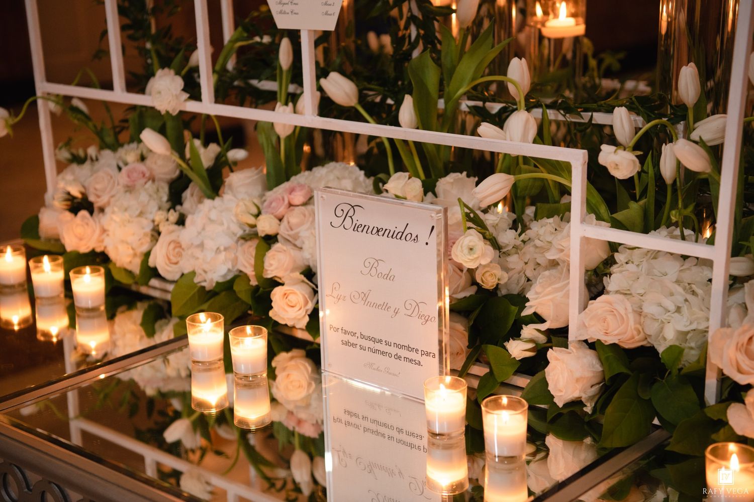 Elegant wedding welcome sign surrounded by white roses, tulips, and glowing candles on a mirrored table.