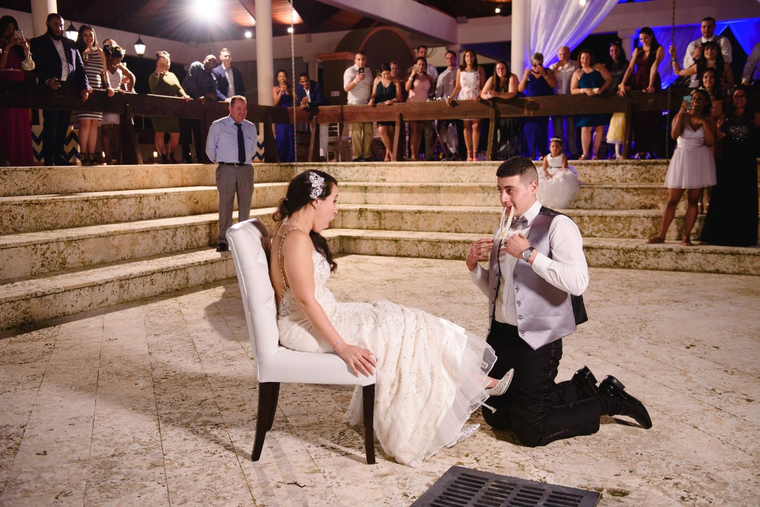 Groom kneeling before bride seated on white chair during wedding reception with guests watching on stairs.
