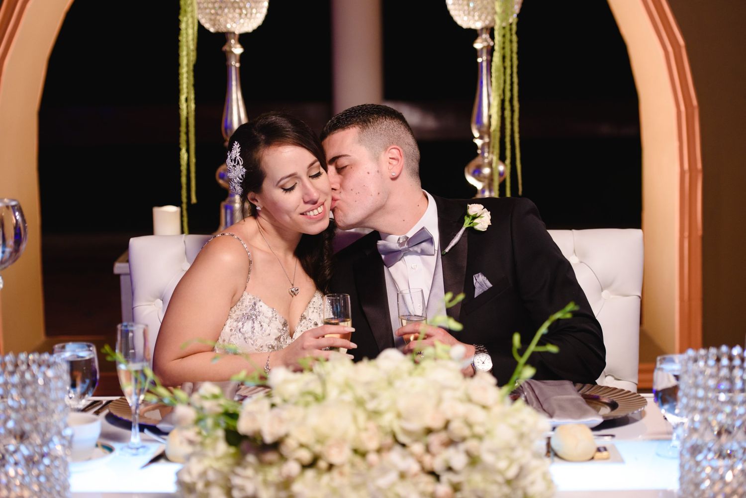 Bride and groom share a romantic kiss at their wedding reception sweetheart table with floral centerpieces.