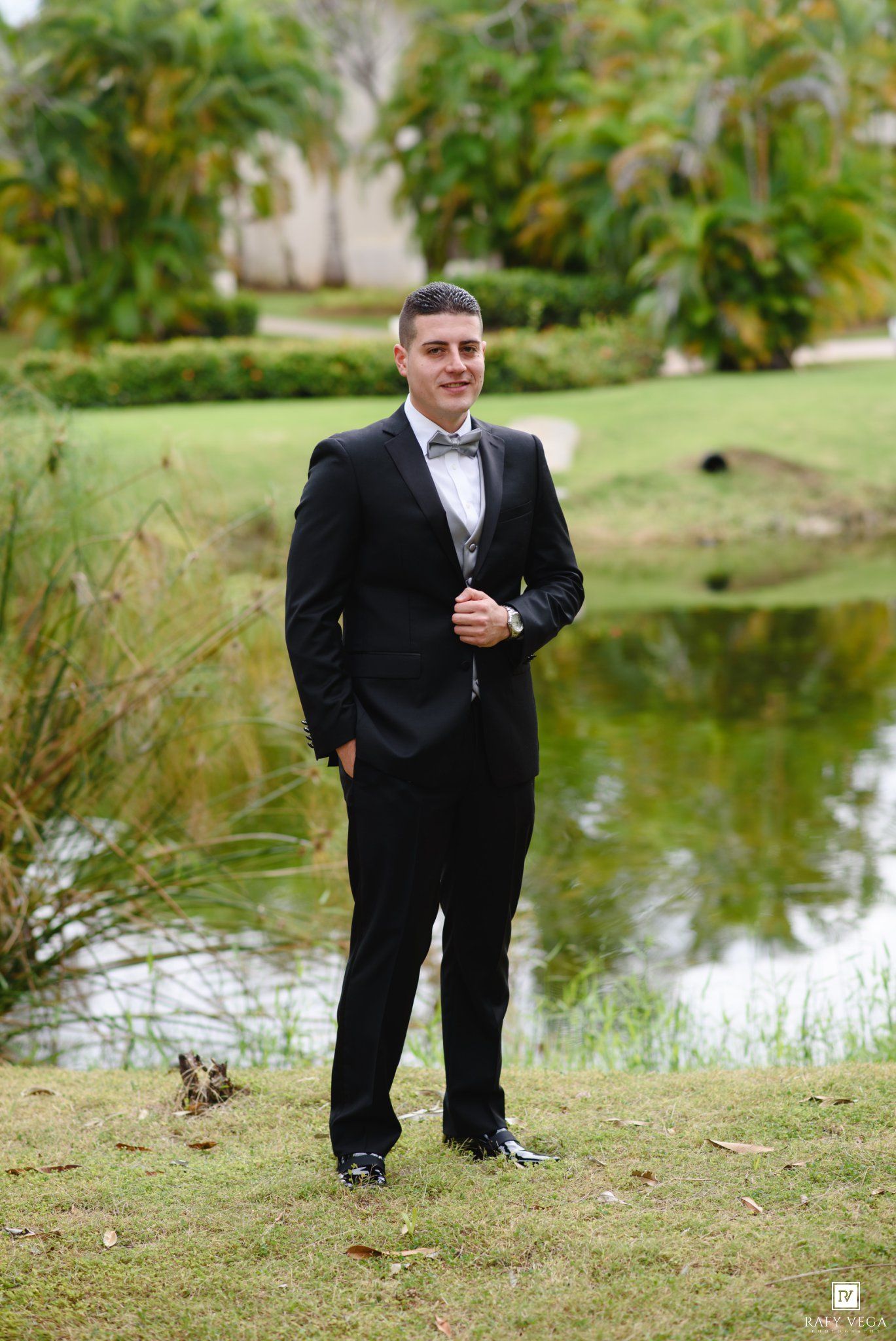 Young man in black tuxedo with bow tie posing outdoors near a pond with lush green tropical landscape.