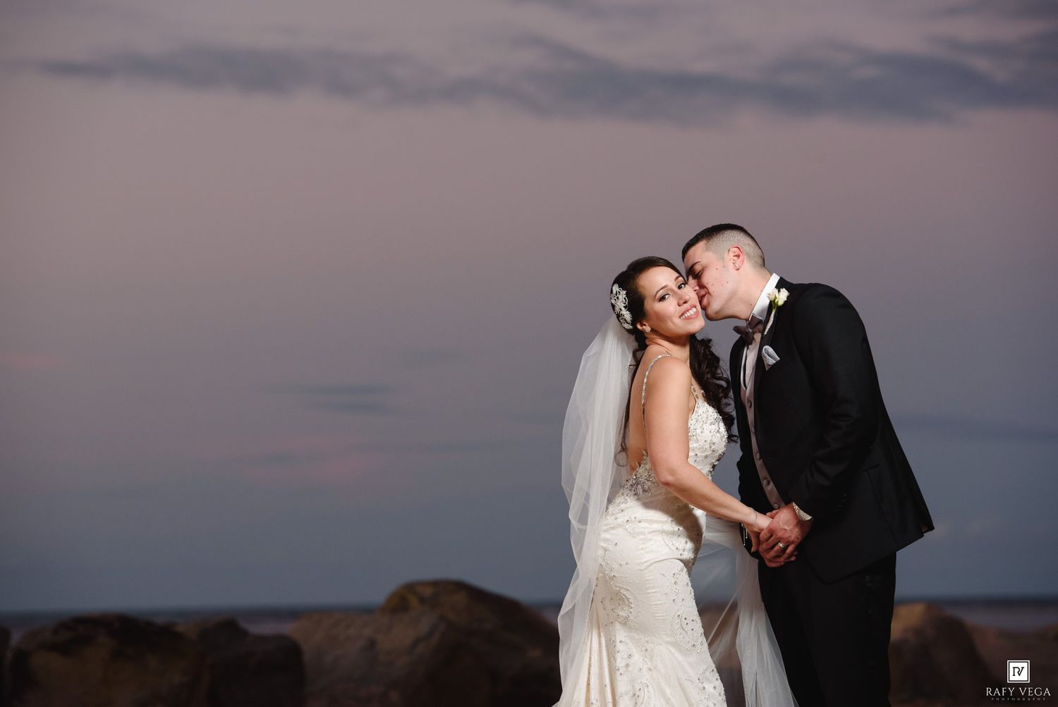 Groom kisses bride on cheek at sunset, she smiles in white gown and veil, rocky shoreline behind them.