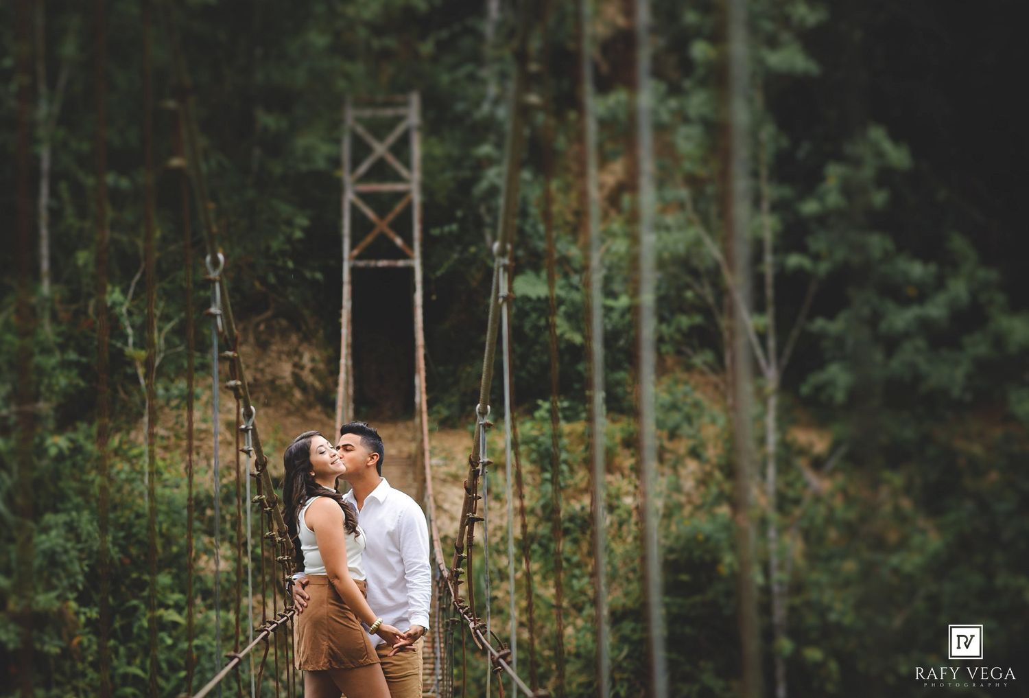 Couple sharing a romantic moment on a suspension bridge surrounded by lush green forest trees.