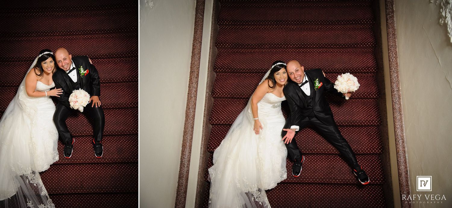Bride and groom posing on red carpeted stairs in wedding attire, bride holding white bouquet, captured from above angle.