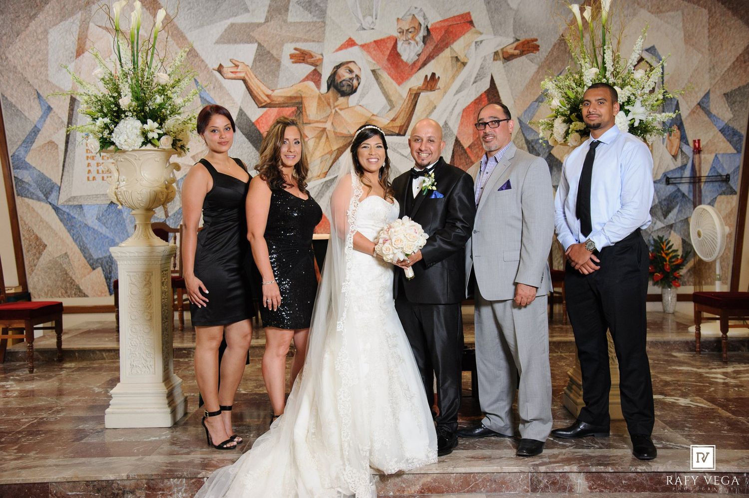Wedding party poses with bride and groom in front of church mosaic after ceremony.
