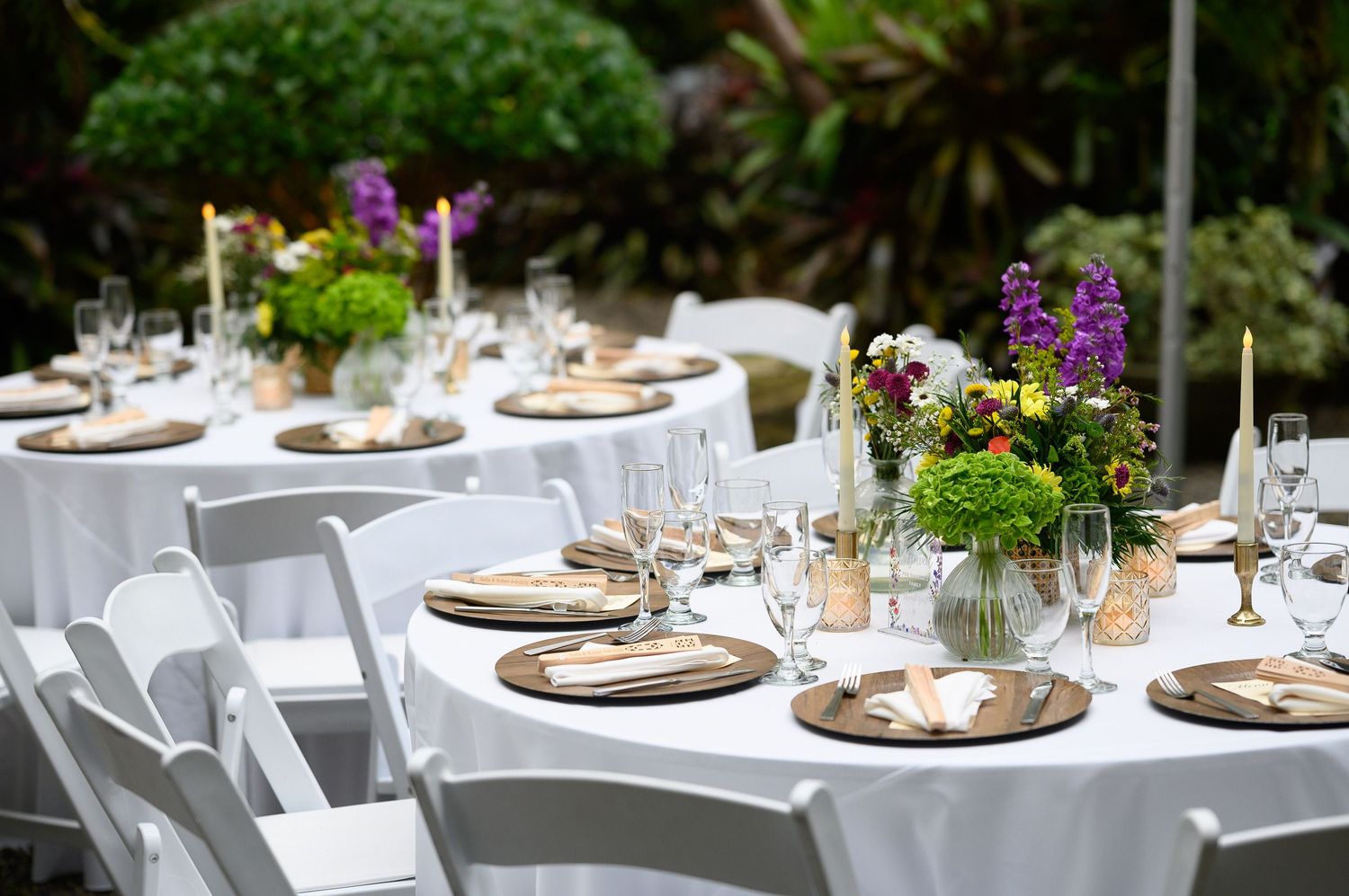 Elegant outdoor wedding reception tables with white linens, wood chargers, colorful floral centerpieces, and glassware.