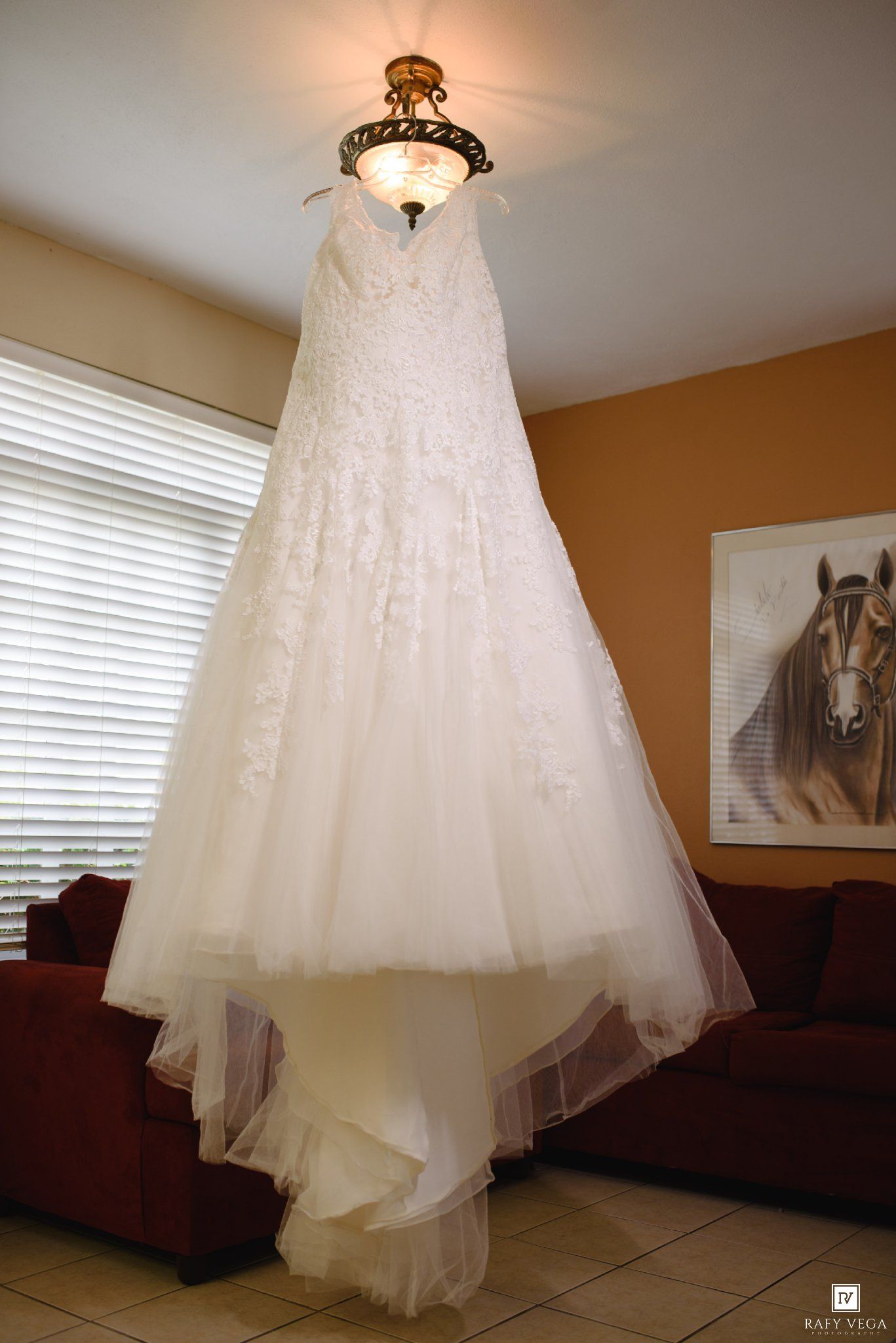 White lace wedding gown hanging from a ceiling light fixture in a warmly decorated room with a horse painting.