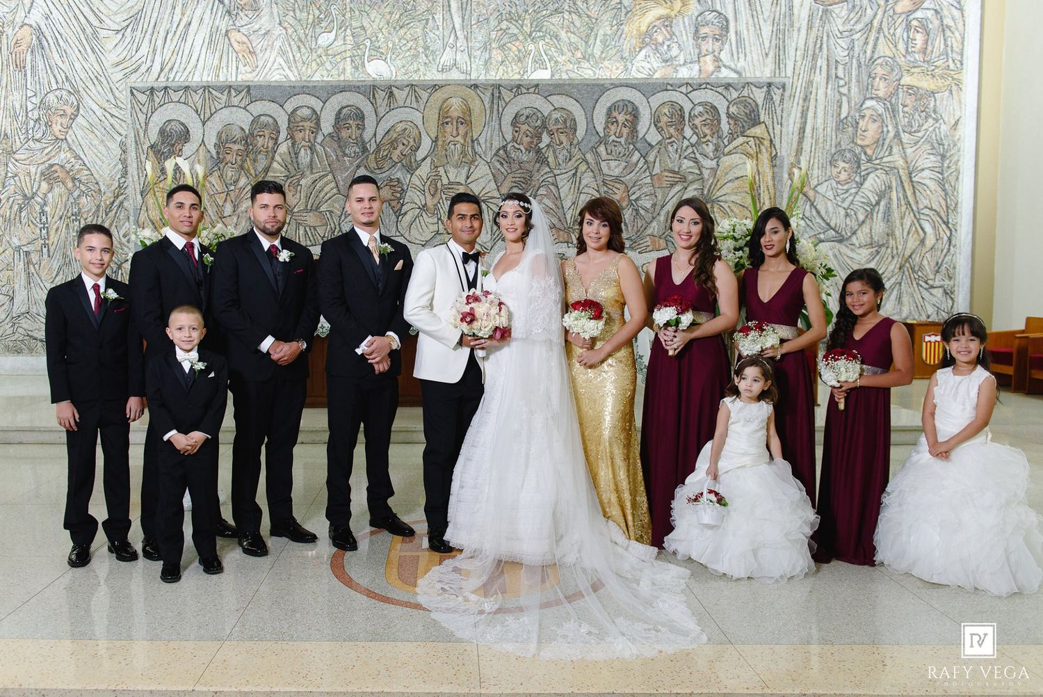 Wedding party poses in church with mosaic backdrop, bride in white gown, groom in white tux, bridesmaids in burgundy and gold.