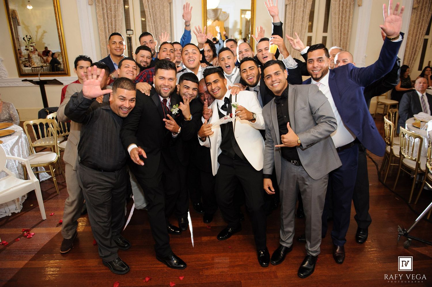 Large group of wedding guests in suits celebrate together, smiling and waving at a elegant reception venue.