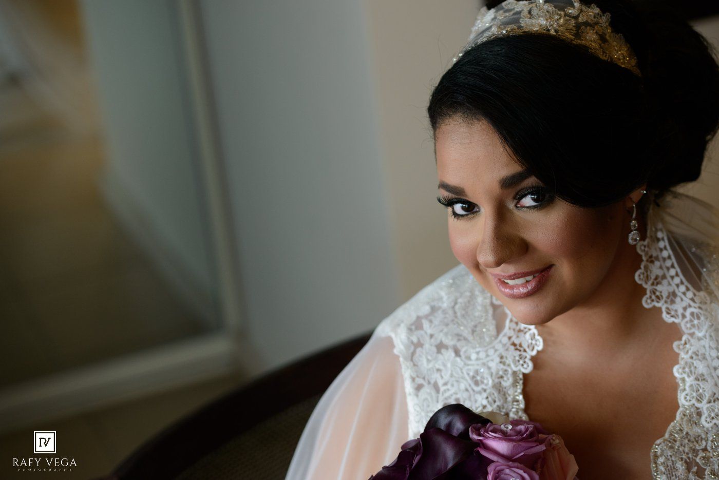 Elegant bride wearing a lace wedding gown and tiara, smiling softly while holding a purple bouquet.