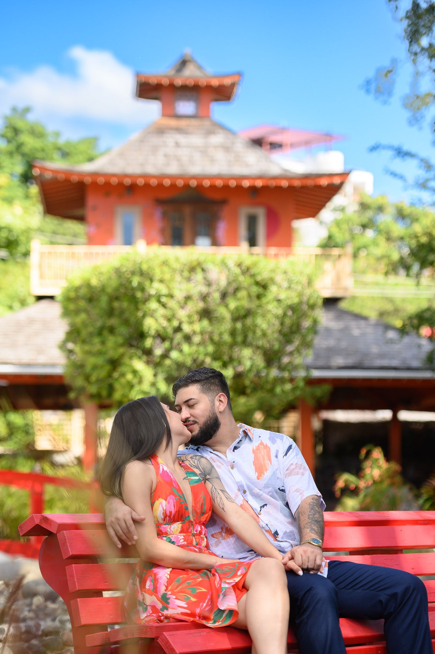 Couple kissing on a red bench in front of a red Asian-style pagoda building surrounded by lush green trees.