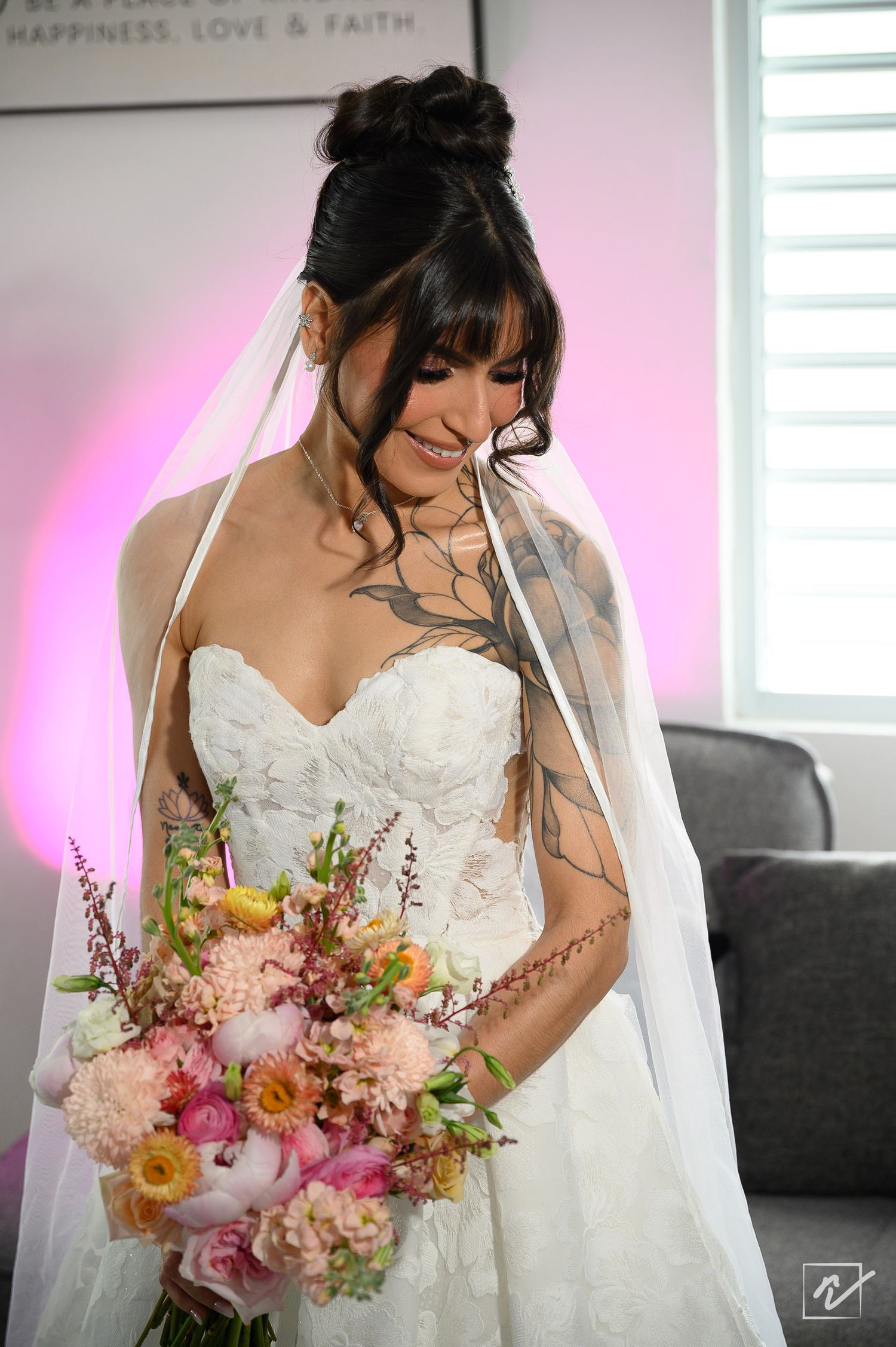 Smiling bride in a strapless white lace gown and long veil holds a colorful pink and peach bouquet while standing in a softly lit room.