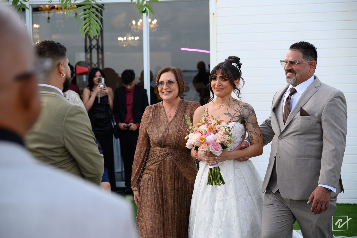 Bride in a white lace gown holding a pink bouquet walks down the aisle beside a man in a gray suit as wedding guests look on.