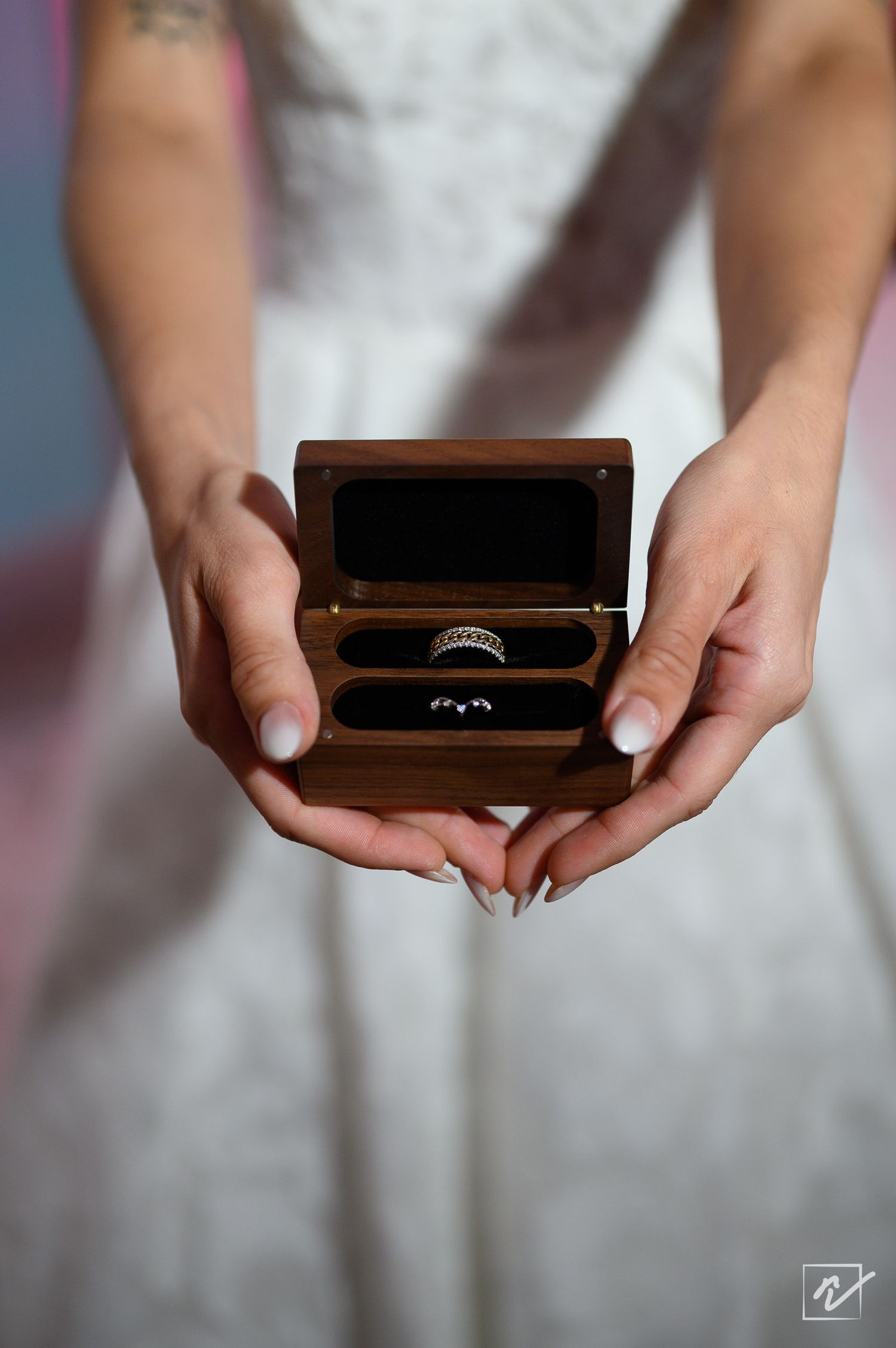 Bride in a white lace gown holds an open wooden ring box displaying two wedding bands against a softly blurred background.