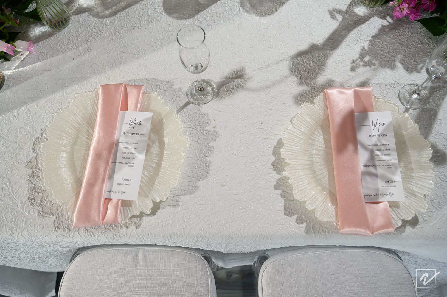 Overhead view of an elegant wedding table with blush pink napkins, printed menu cards, clear charger plates, and white chairs on lace linen.
