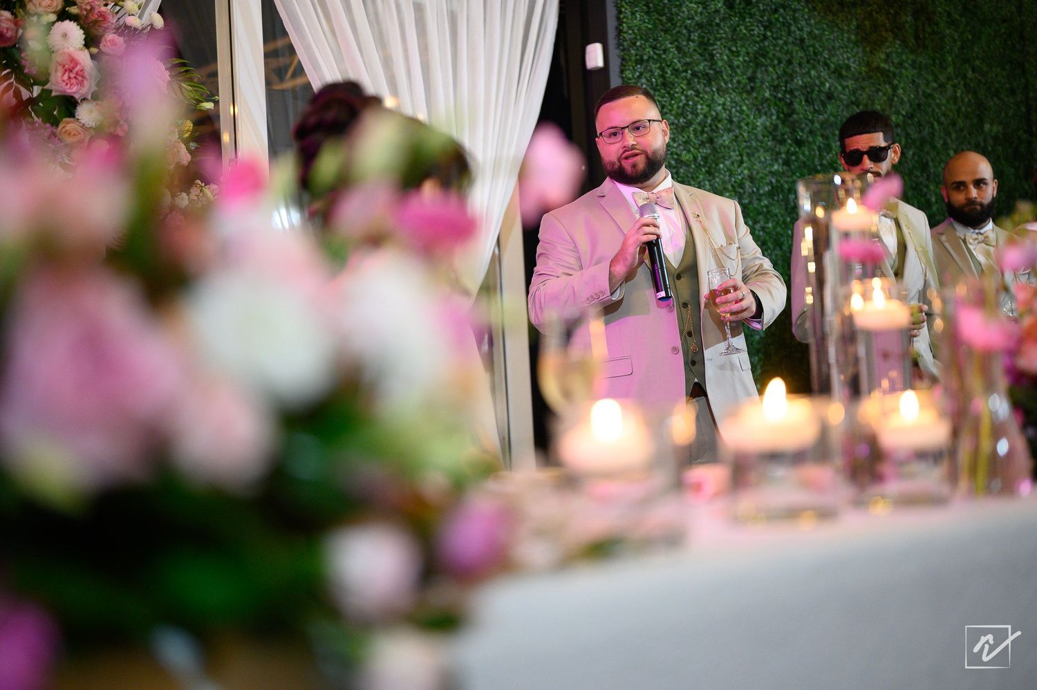 Bearded man in a light suit speaks into a microphone at a candlelit wedding reception with pink flowers and guests seated nearby.