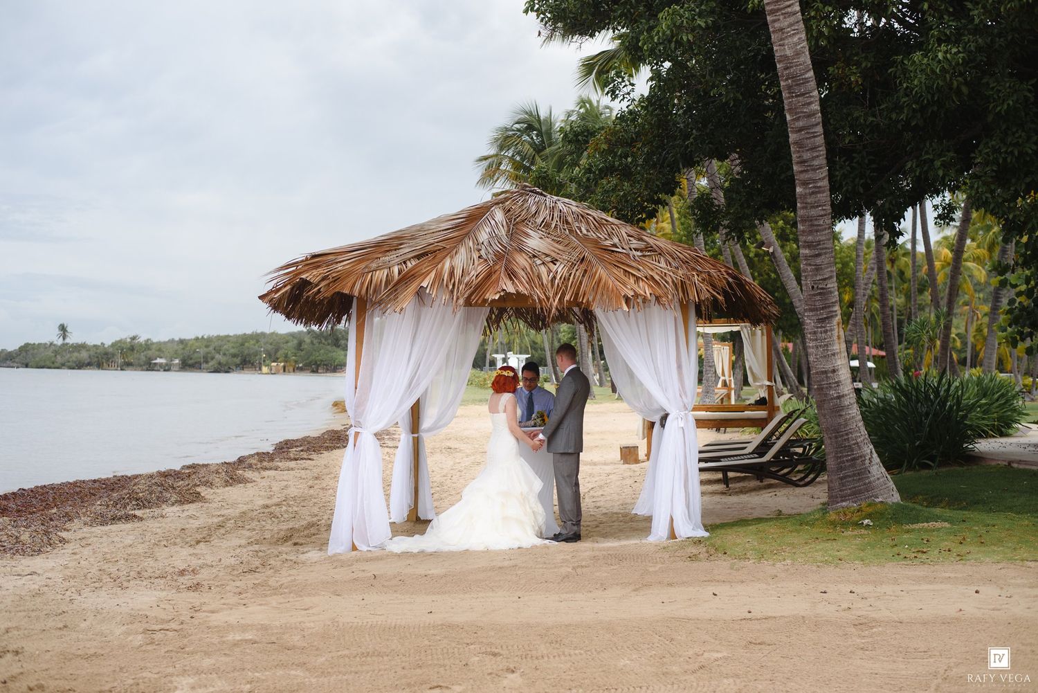 Couple exchanging wedding vows under a thatched gazebo with white drapes on a tropical beach.