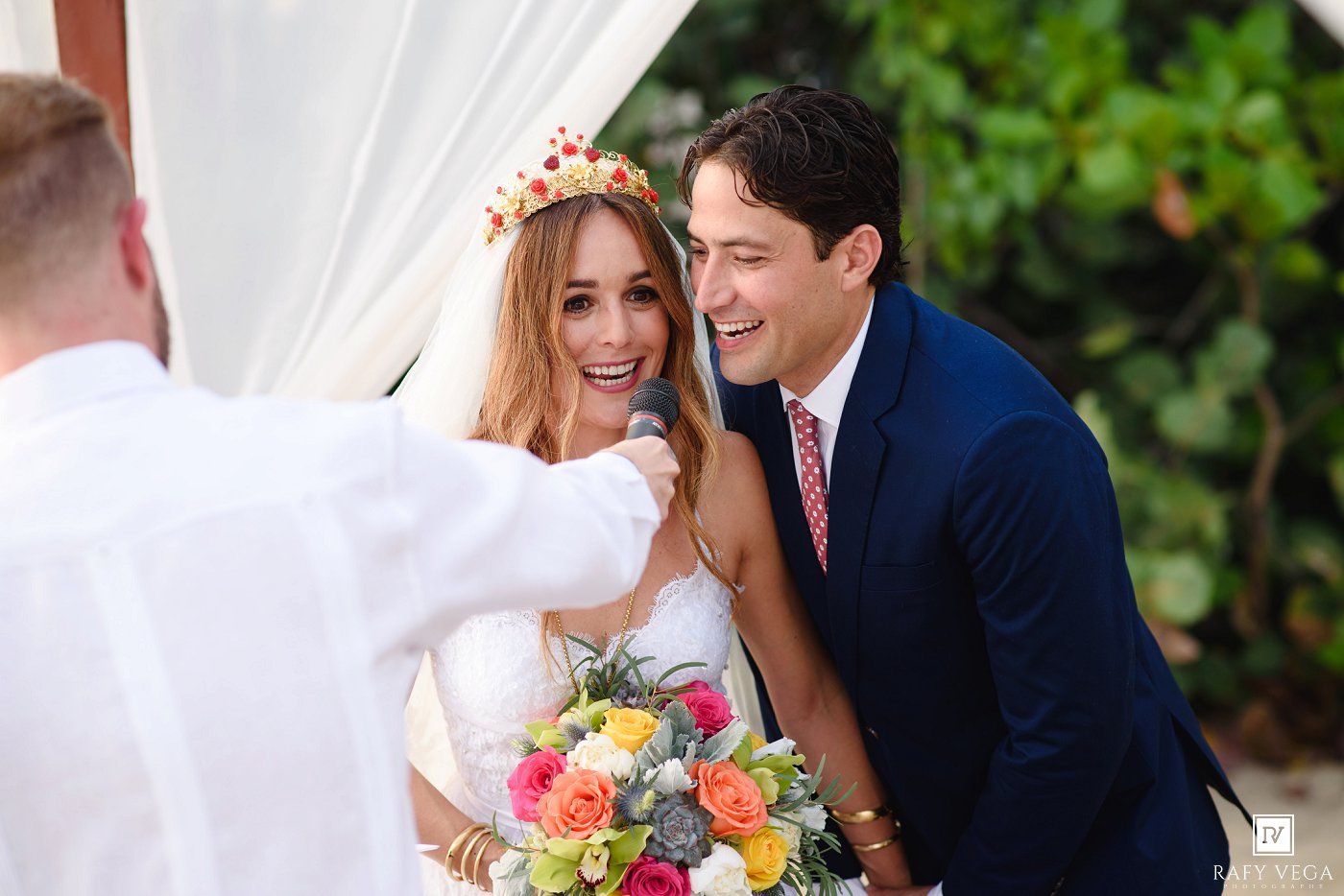 Bride with floral crown and groom in navy suit laughing during outdoor wedding ceremony with colorful bouquet.