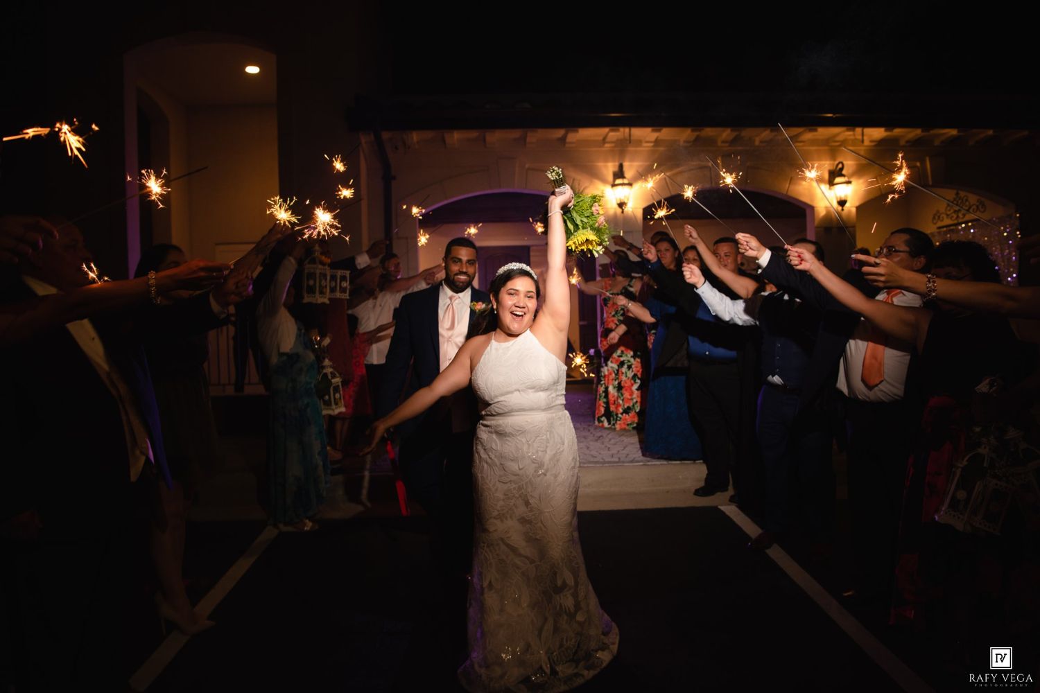 Bride and groom walk through sparkler exit tunnel surrounded by cheering wedding guests at night.