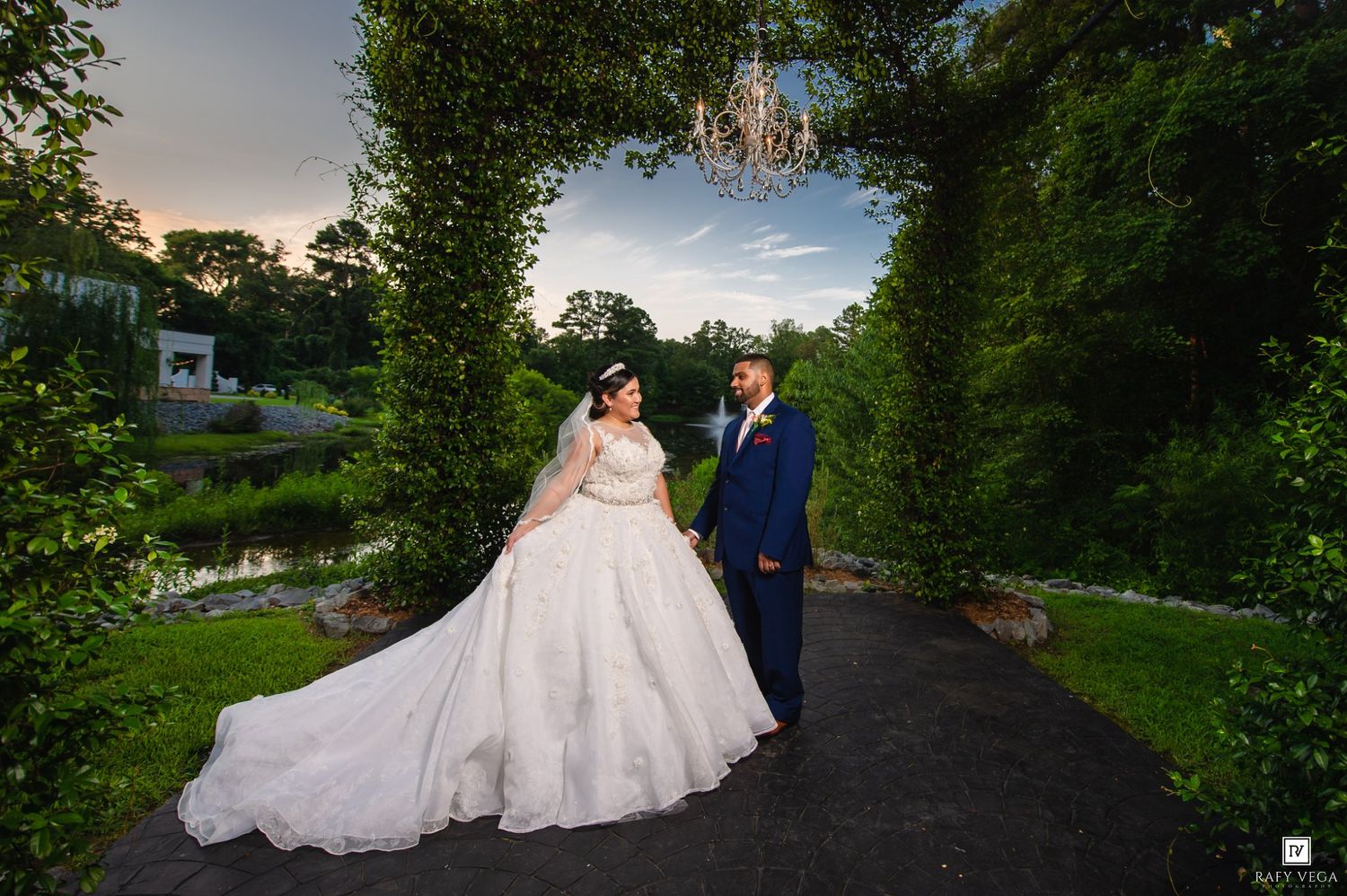 Bride in ball gown and groom in navy suit pose on garden path under floral arch at golden hour wedding.