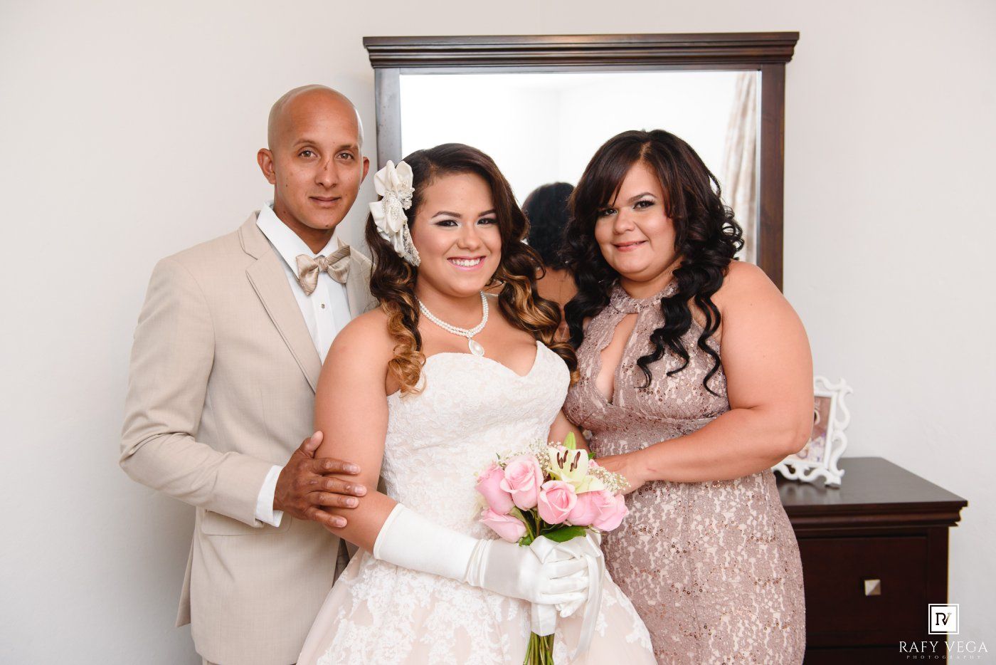 Bride in white gown holds pink bouquet, flanked by groom in tan suit and bridesmaid in pink dress, posing before mirror.