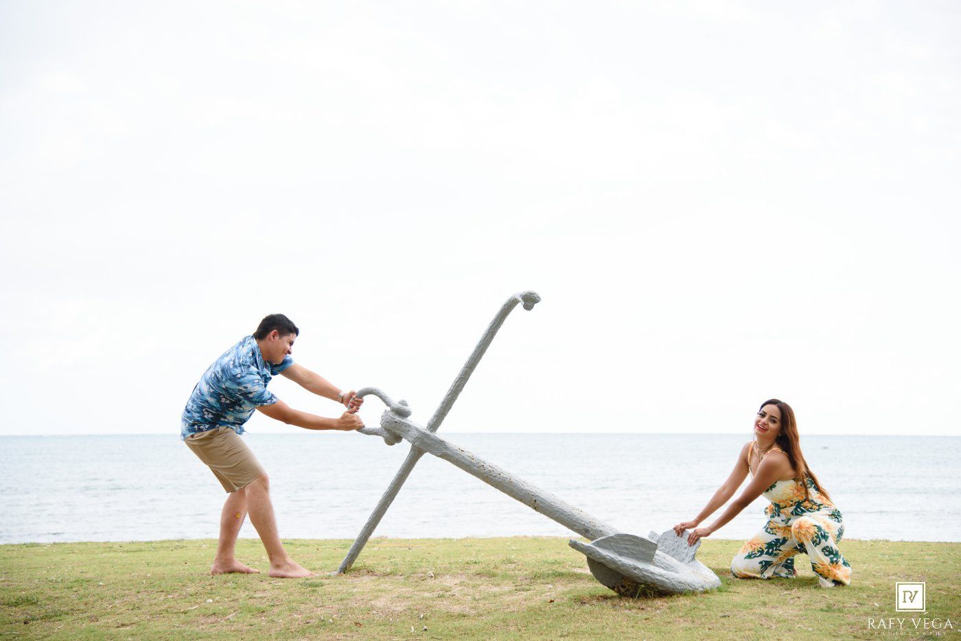 Couple playfully posing with a large anchor on a grassy beachfront, man pushing while woman pulls near the ocean.