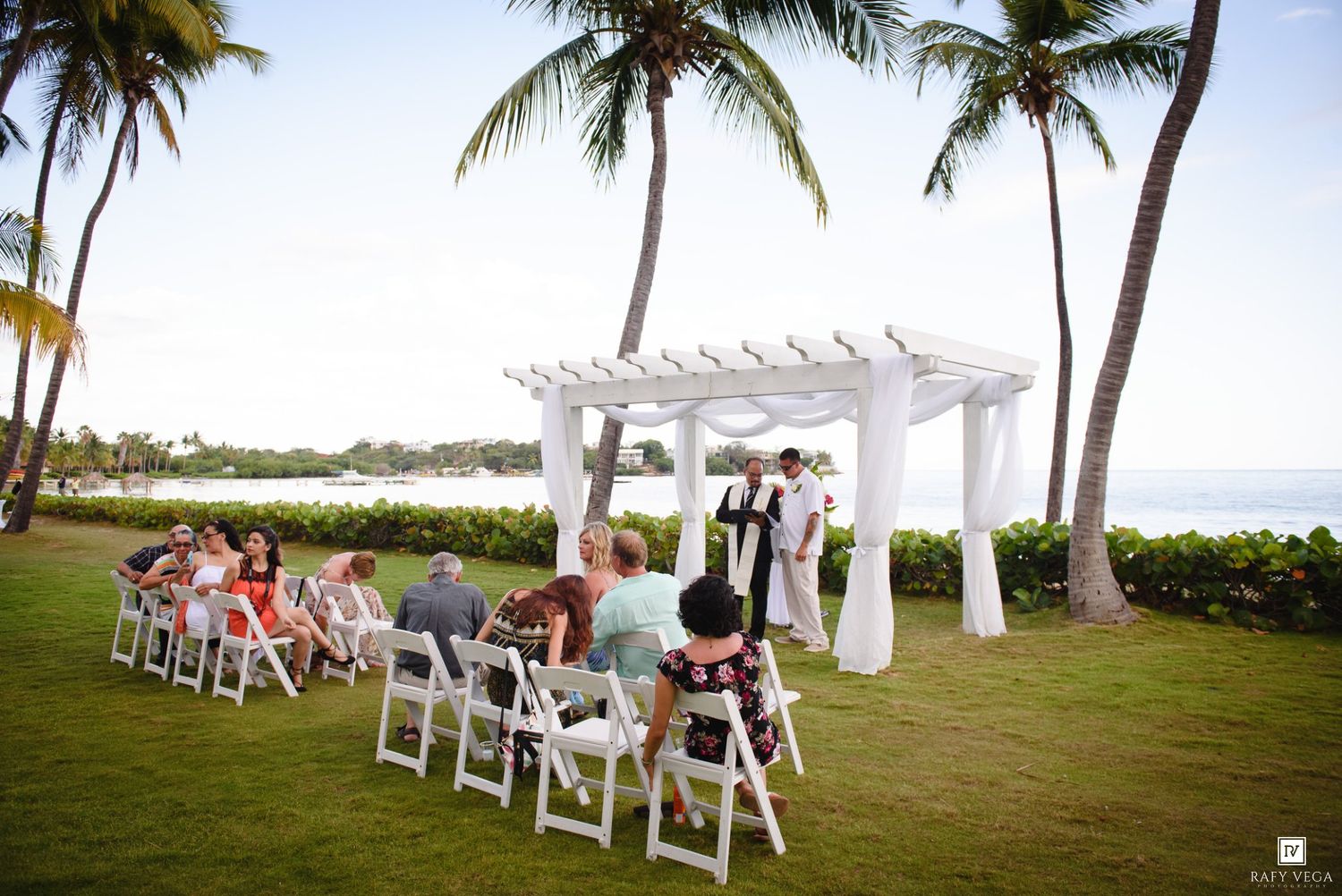 Small intimate outdoor wedding ceremony on a tropical lawn with white chairs and palm trees.