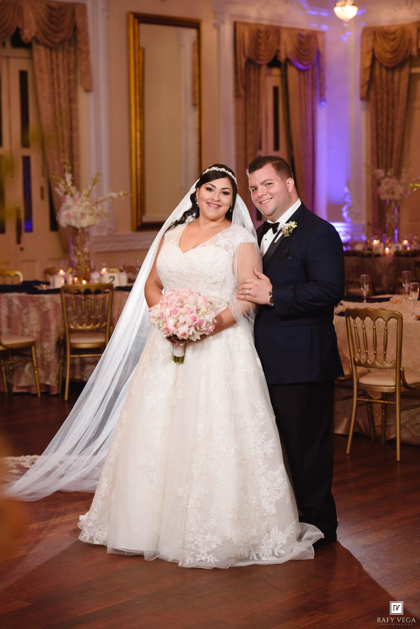 Bride in white lace ball gown with veil and groom in navy tuxedo posing in elegant wedding reception hall.