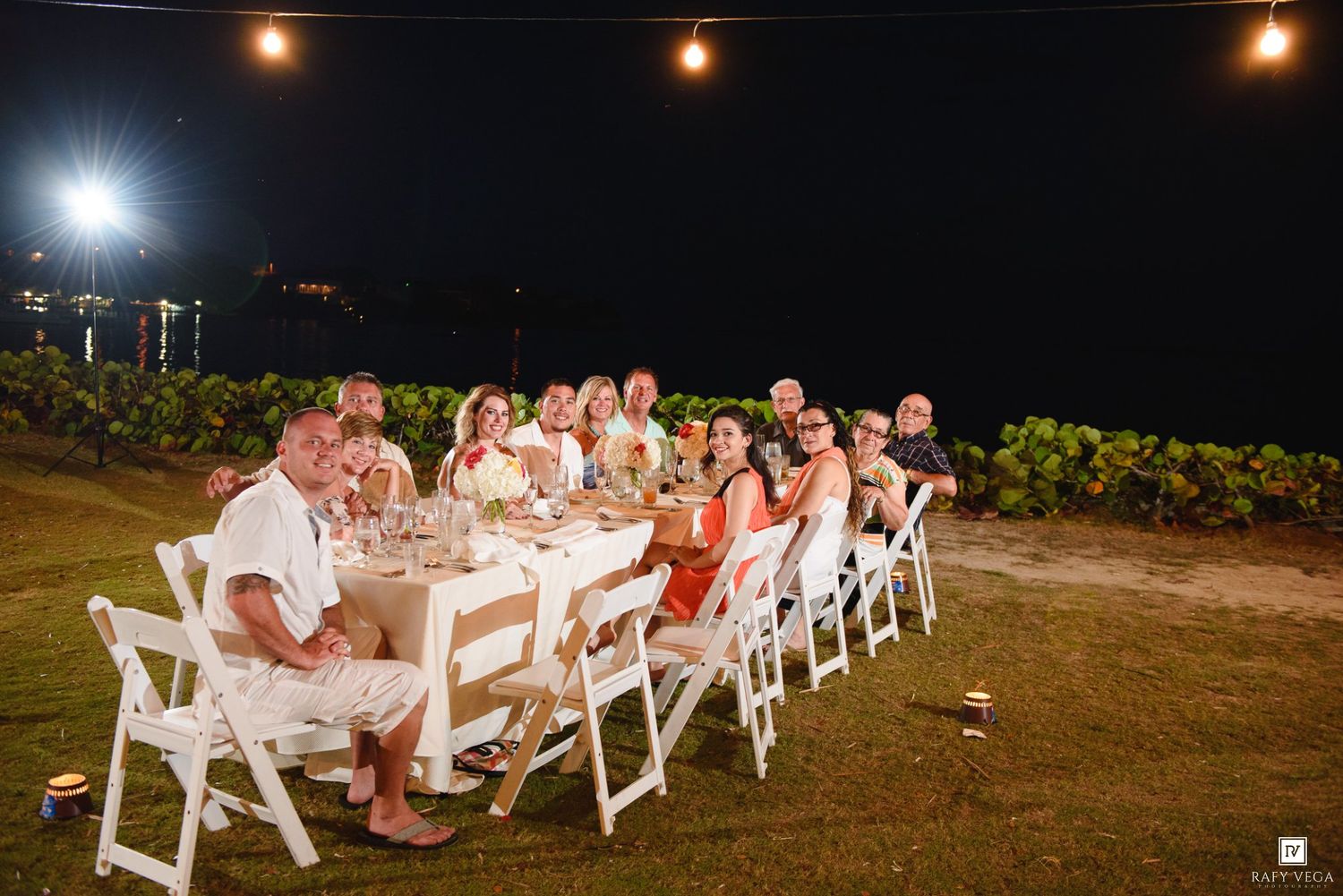 Group of guests dining at an outdoor evening reception table with string lights in a tropical garden setting.