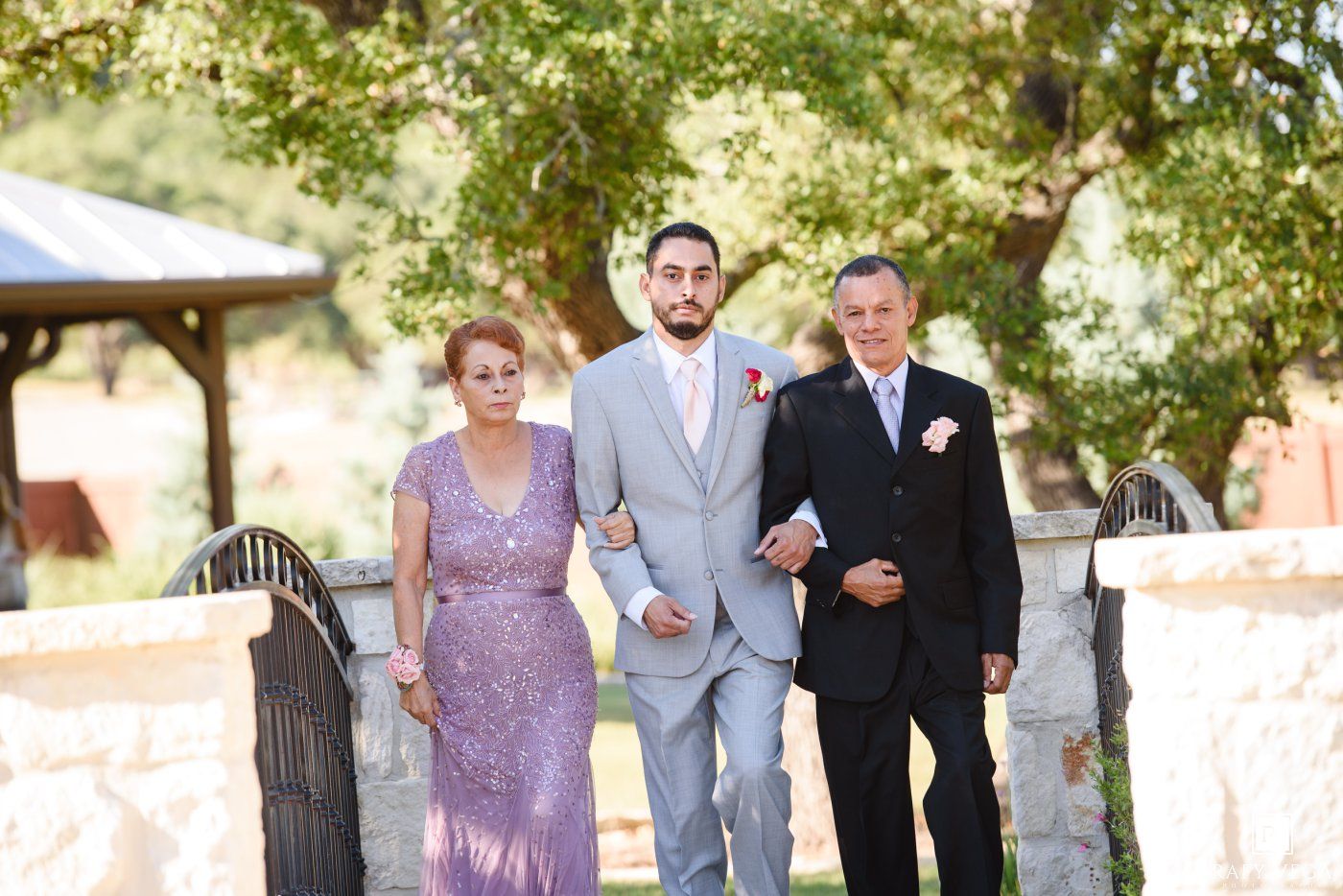 Groom in gray suit walks with parents at outdoor wedding ceremony surrounded by lush green trees.